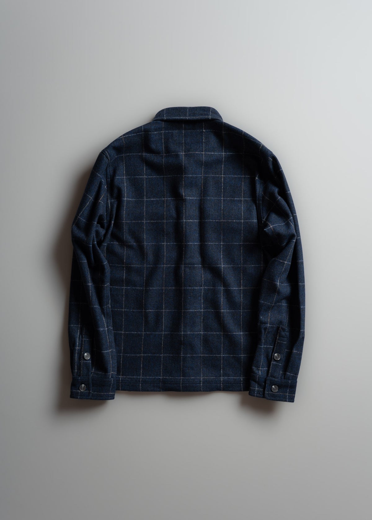 A navy blue plaid shirt with buttoned cuffs is laid flat on a light gray background, showcasing its back design and pattern.