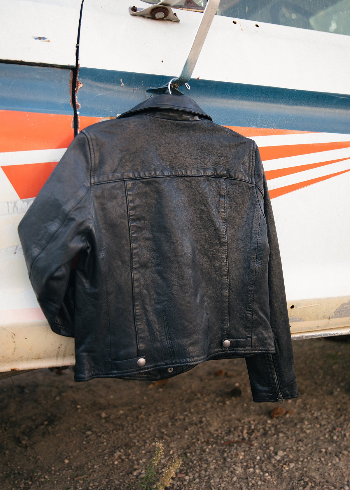 Black leather jacket hangs on a vintage car door with orange stripes, set against a gravel background.
