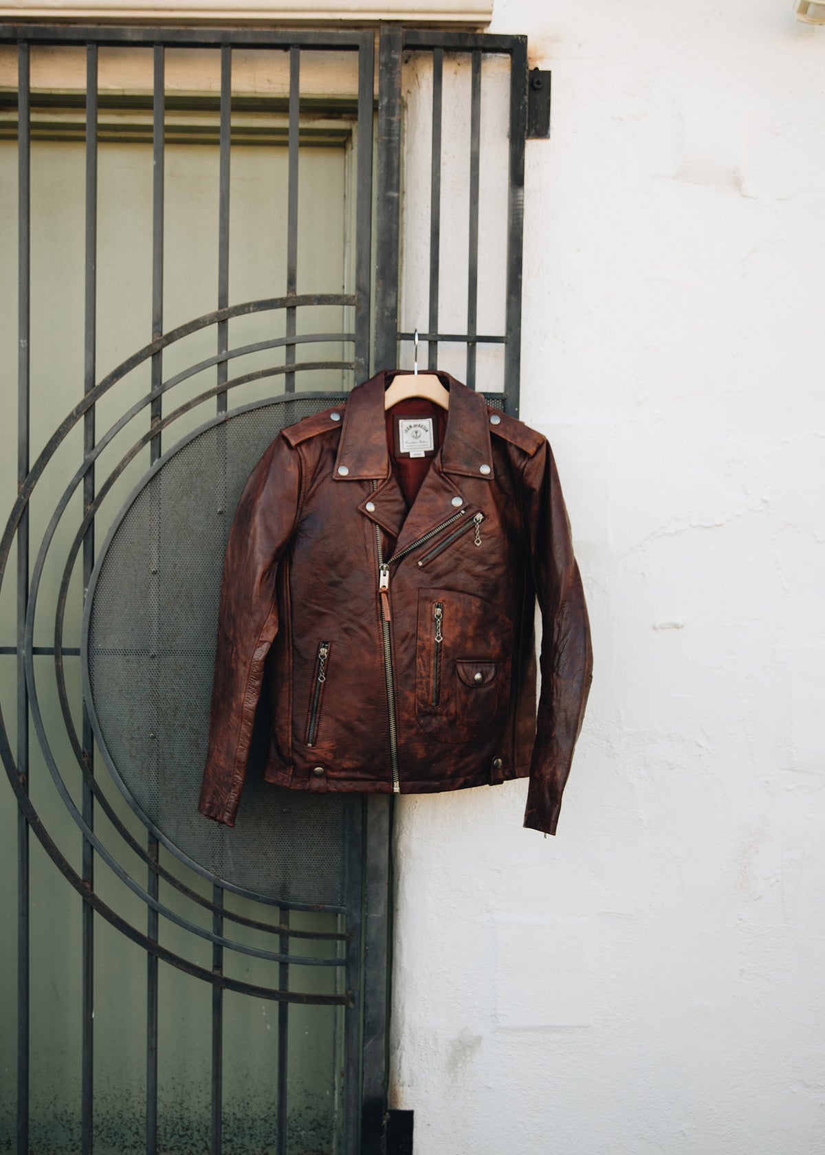 Brown leather jacket hangs on a metal gate against a white wall, showcasing its vintage style and detailed zippers.