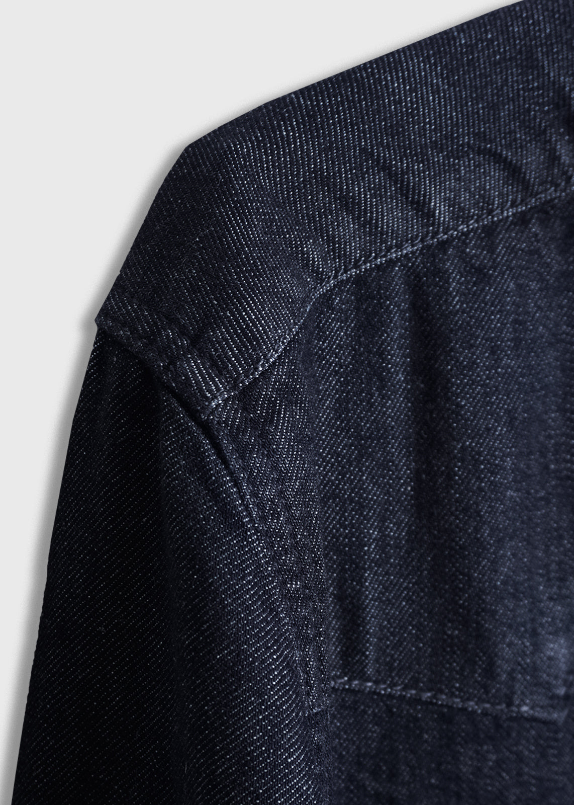 Close-up of a dark denim jacket's shoulder seam, showcasing detailed stitching and texture against a light gray background.