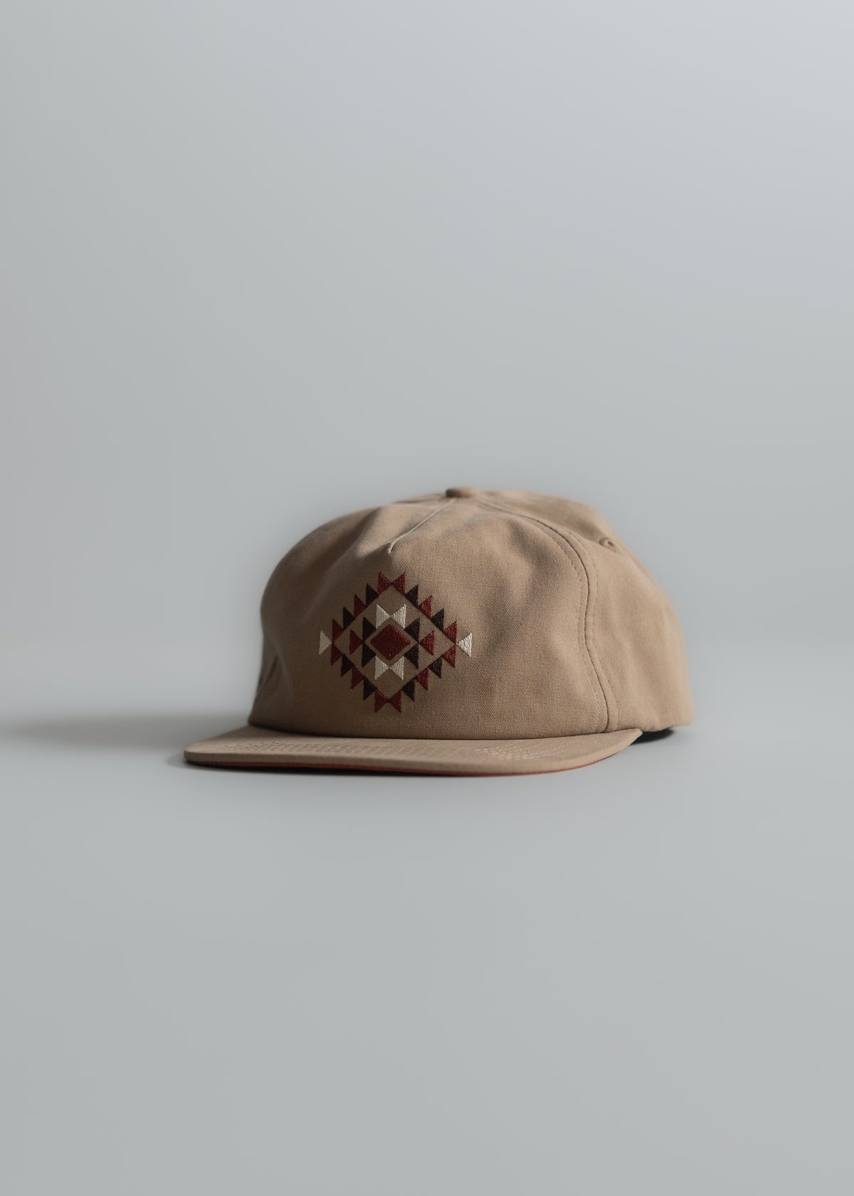 Tan cap with geometric pattern on front, set against a plain gray background. The design features red and white diamond shapes.