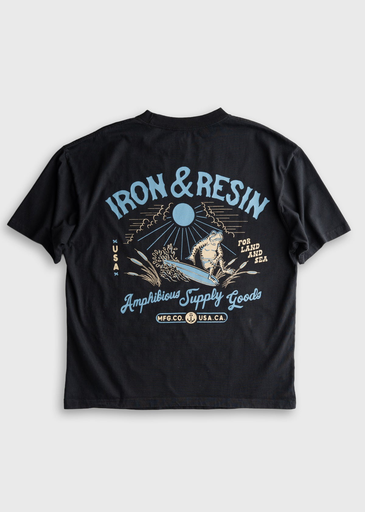 Black T-shirt with "Iron & Resin" logo, featuring a surfer and wave graphic. Text reads "Amphibious Supply Goods, USA."