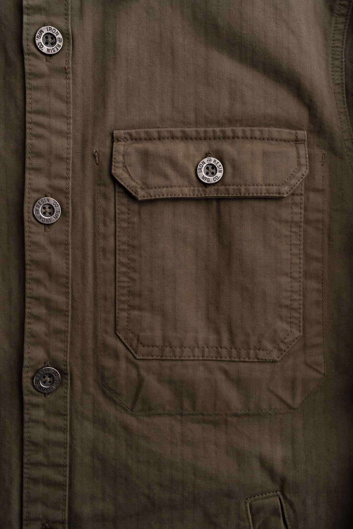 Close-up of a brown jacket with a buttoned flap pocket. Buttons are engraved with "Iron and Resin MFG. CO" for a rugged look.