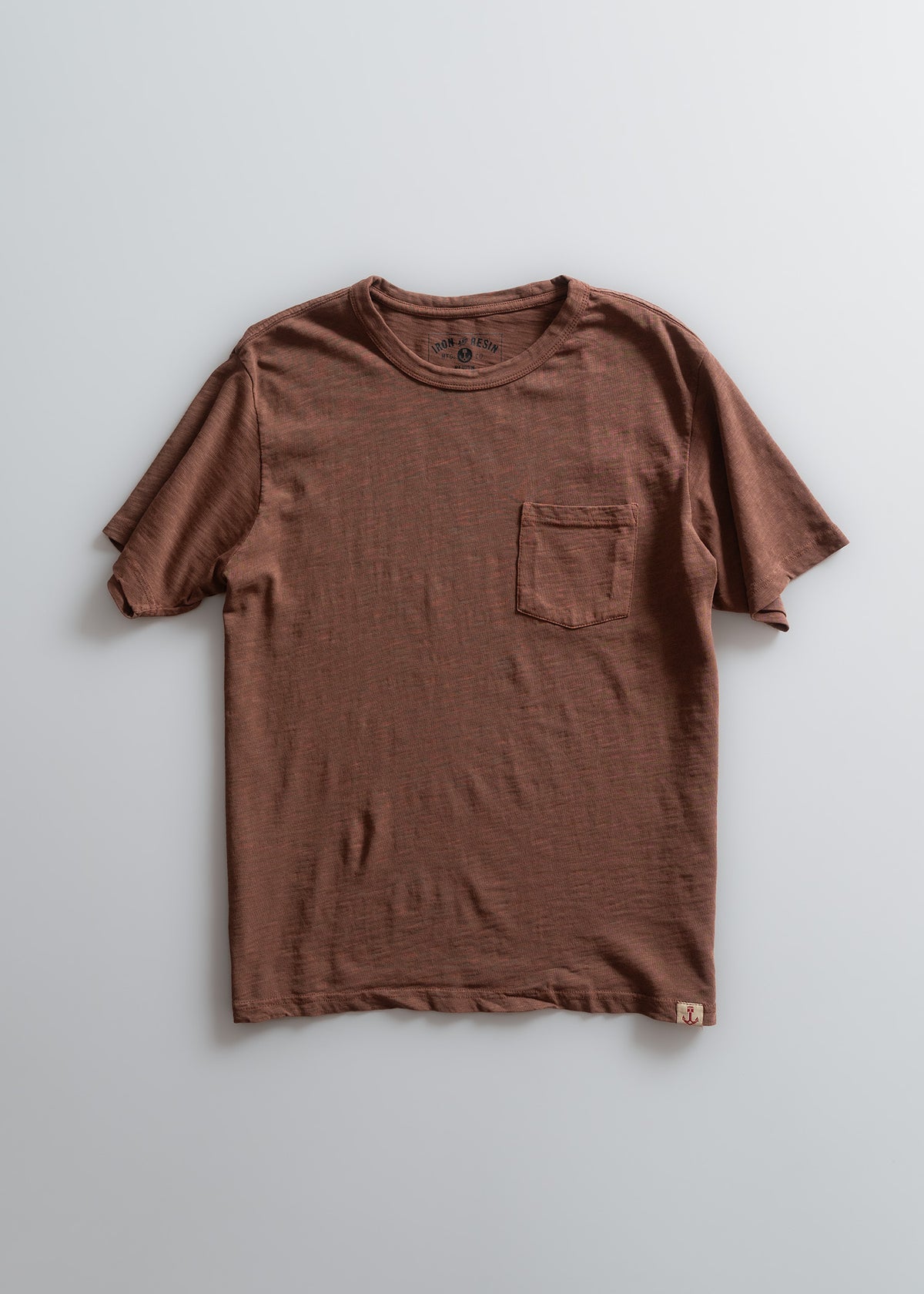 Brown T-shirt with a chest pocket, laid flat on a light background. Simple design, short sleeves, casual style.