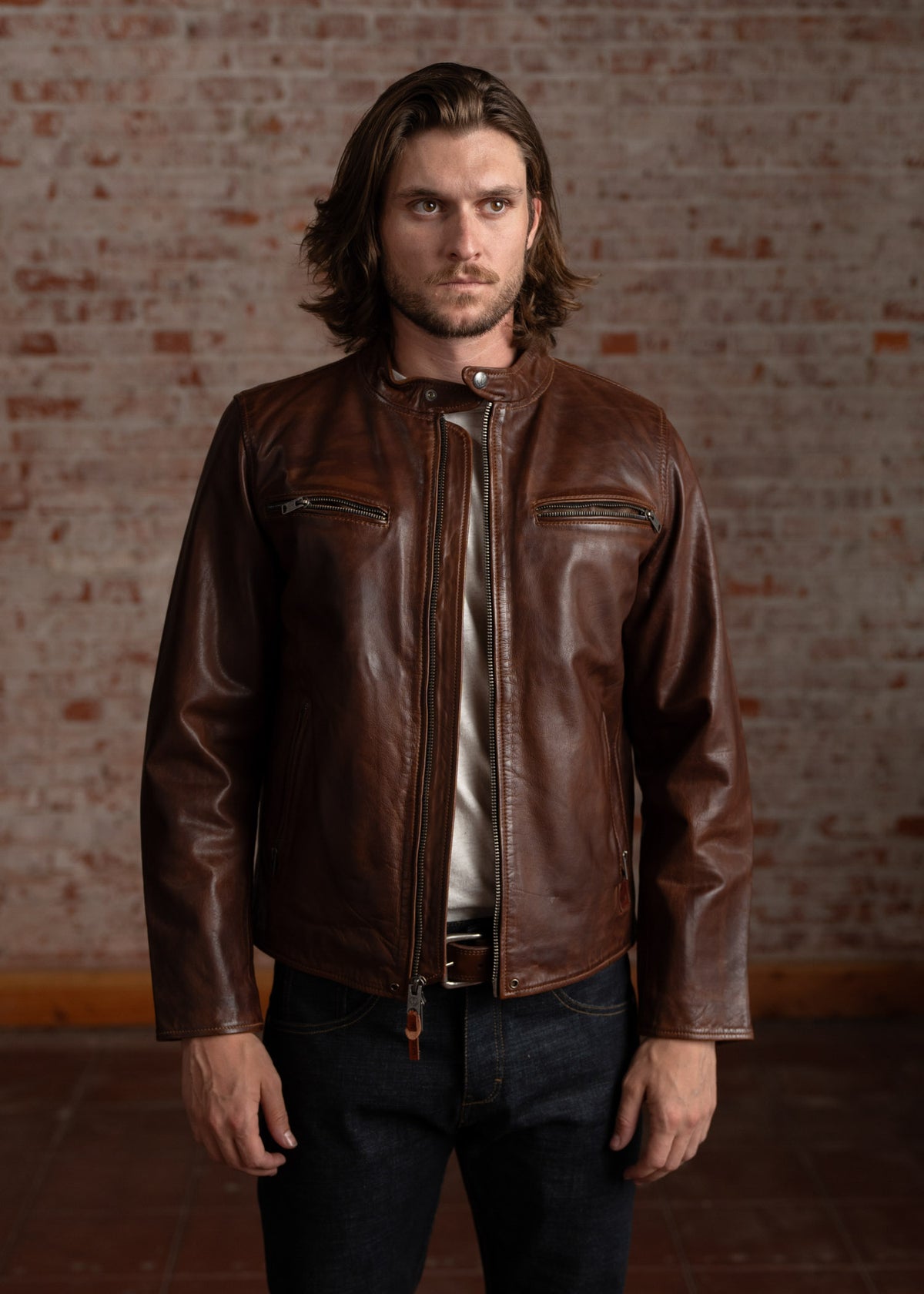 Man in brown leather jacket stands against a brick wall, looking serious. He wears dark jeans and a white shirt underneath.