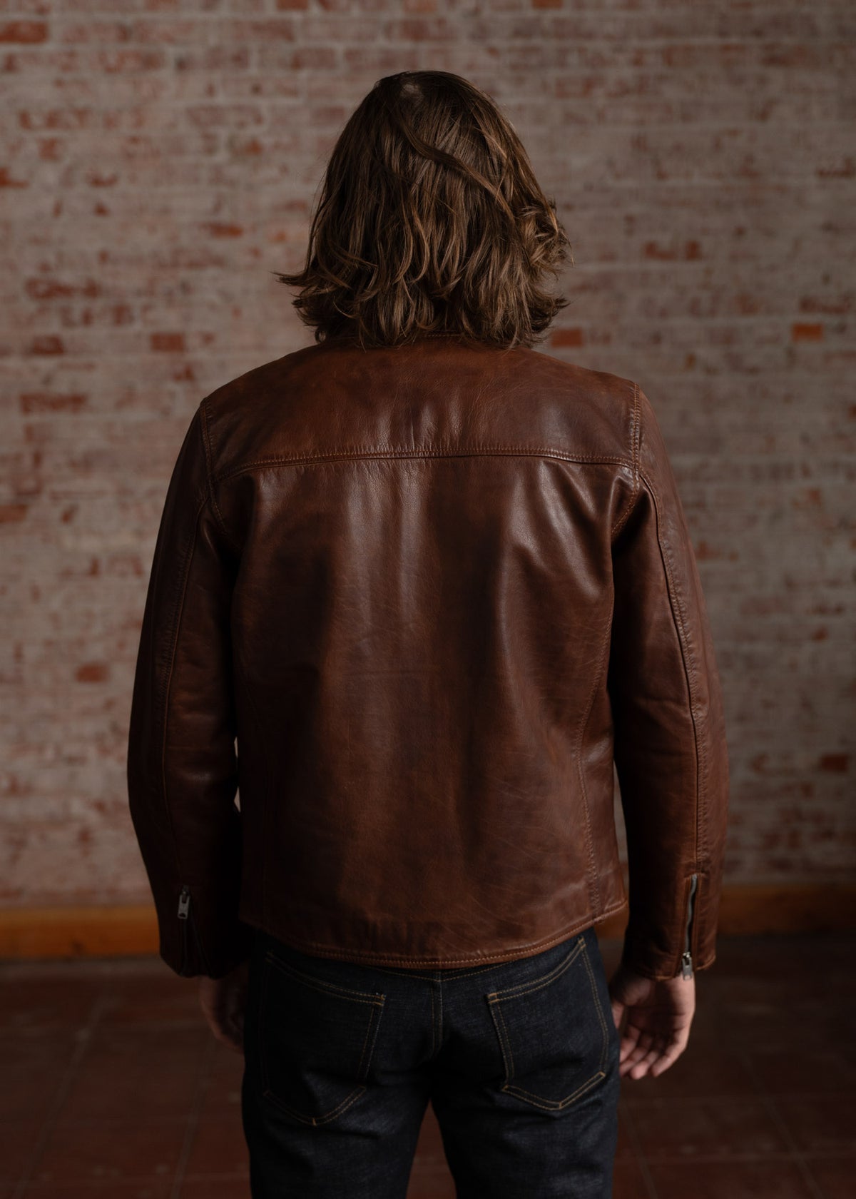 A person with long hair wears a brown leather jacket, facing a brick wall. Denim jeans complete the casual look.