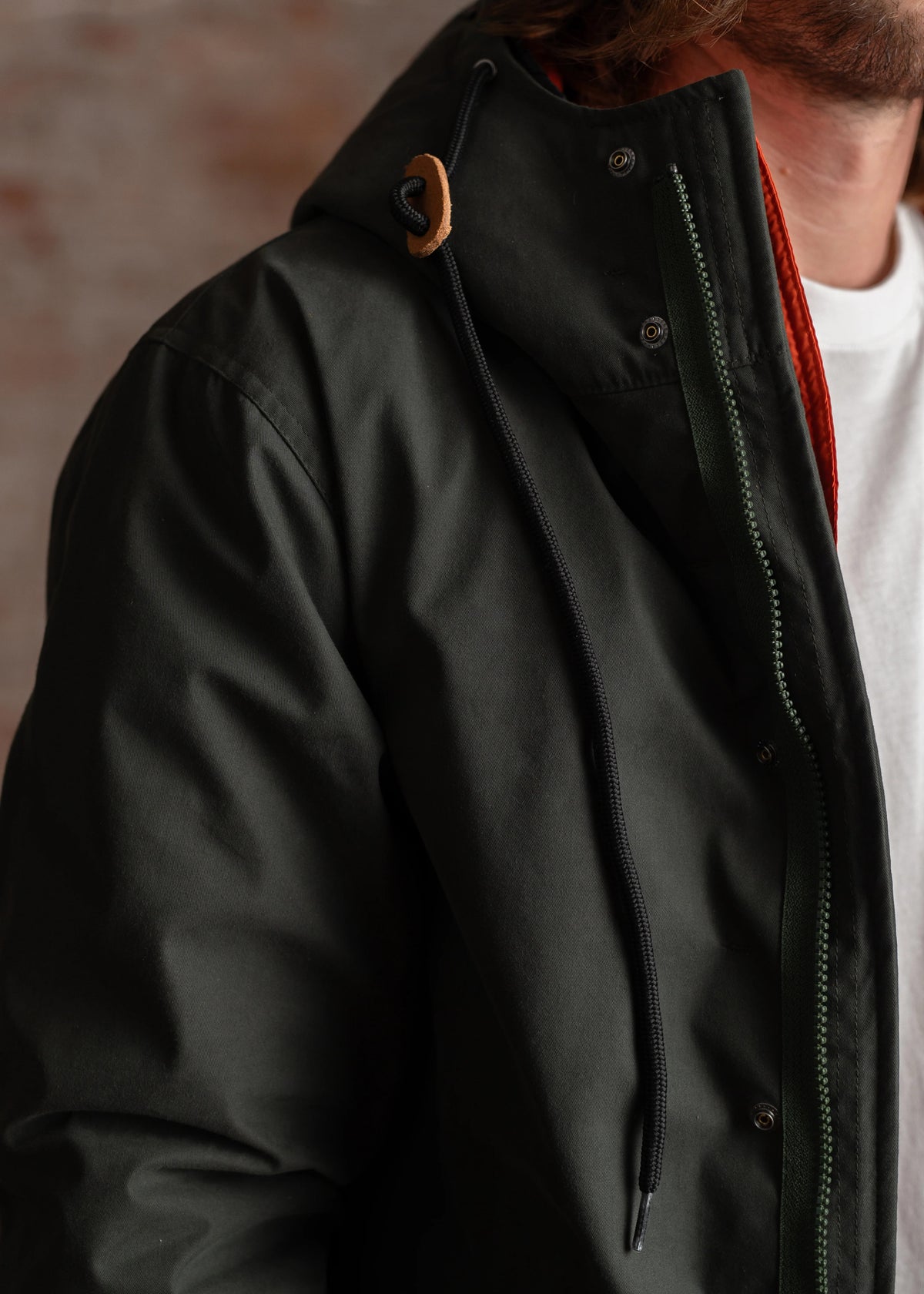 Close-up of a person wearing a dark green jacket with a hood, orange lining, and black drawstring, over a white shirt.