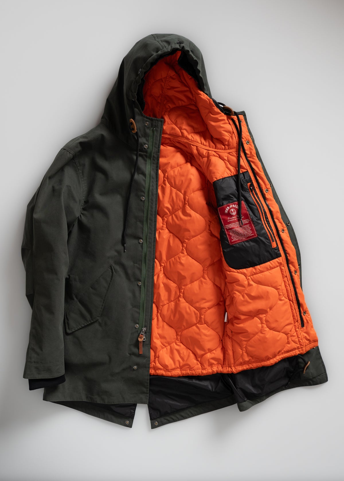 Green jacket with orange quilted lining and hood, open to show interior pocket with red label. Stylish and functional design.