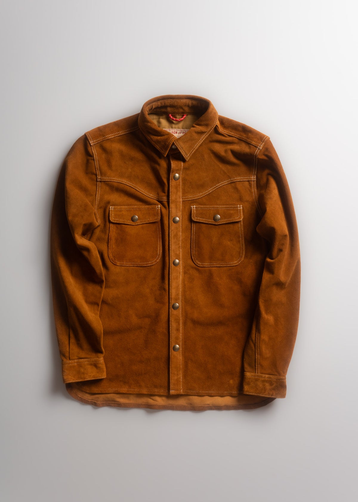 Brown suede shirt with buttoned front and two chest pockets, laid flat on a light surface, showcasing its texture and design.