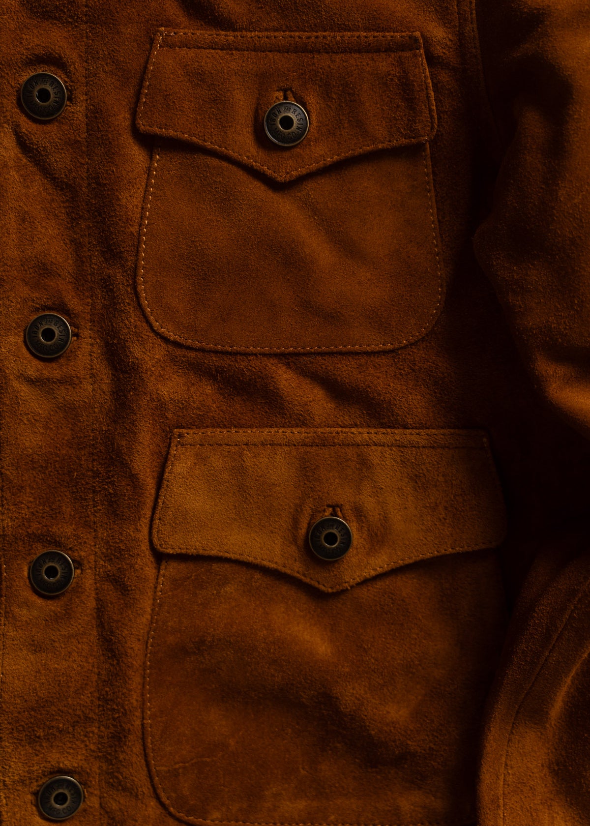 Close-up of a brown suede jacket with two buttoned pockets, highlighting the texture and craftsmanship of the fabric.