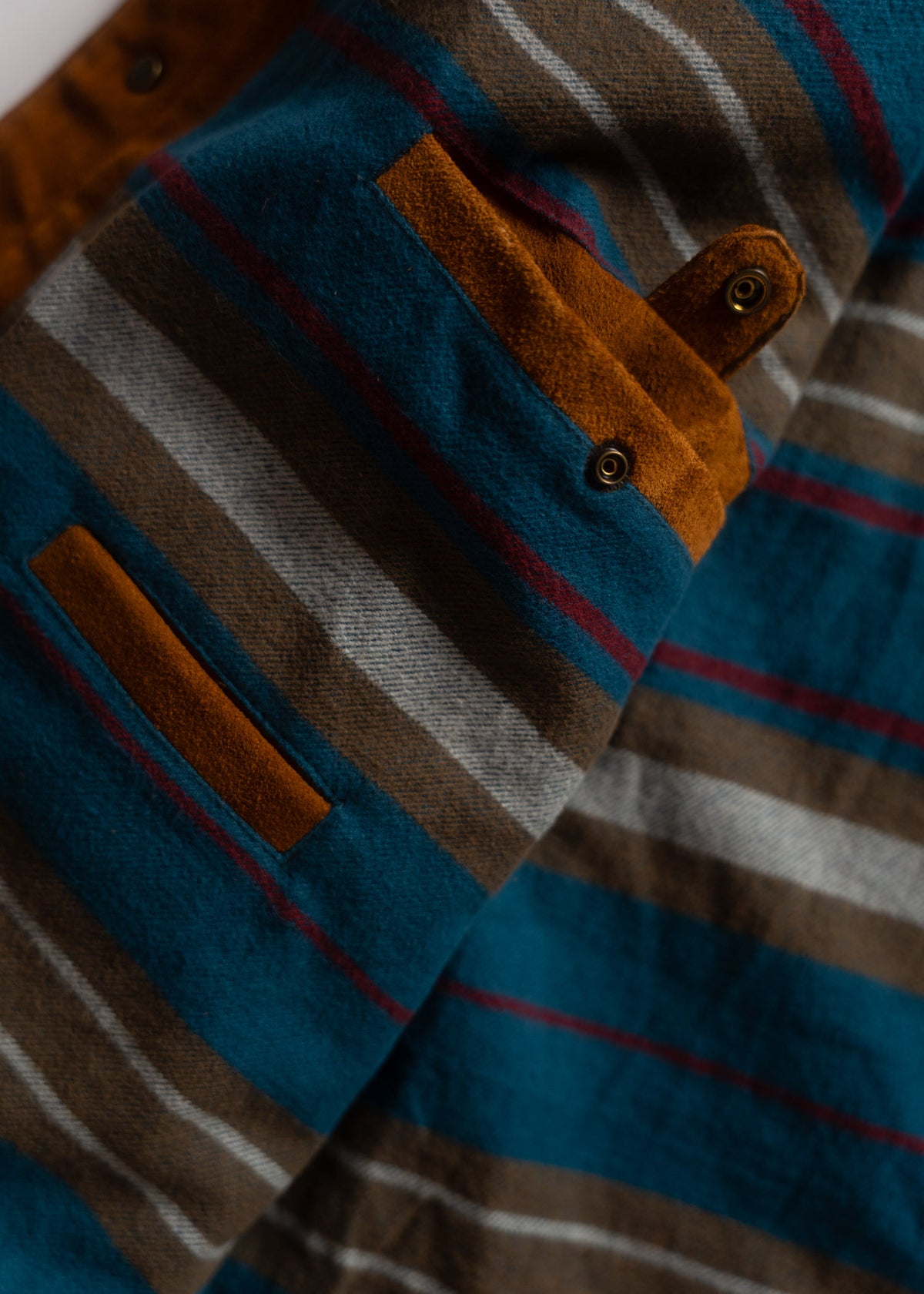 Close-up of a striped fabric with blue, brown, and gray tones, featuring a brown suede pocket and button detail.