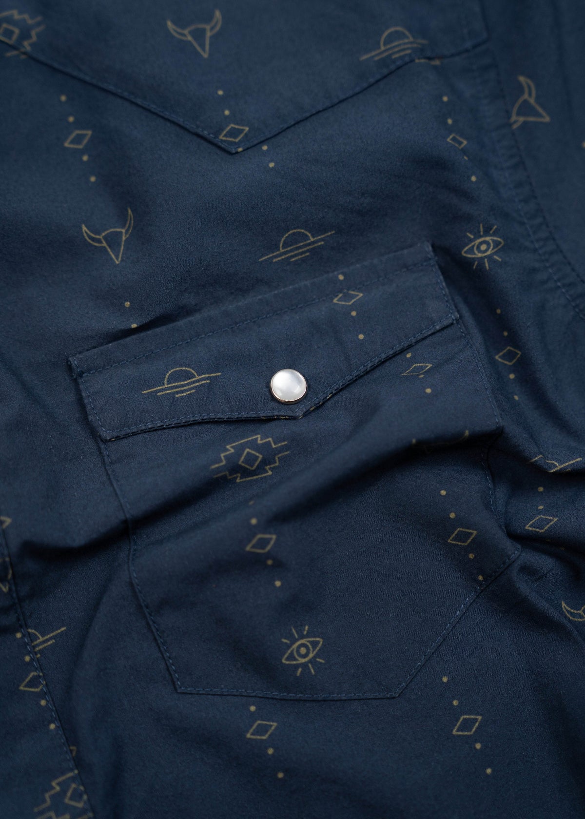Navy shirt with geometric and eye patterns, featuring a silver button on the pocket. Subtle designs add a stylish touch.