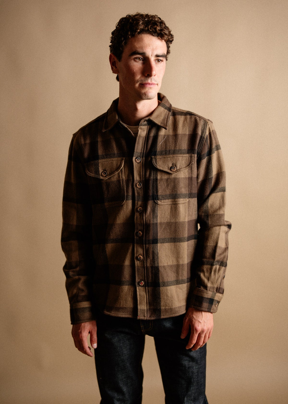Man in a brown plaid shirt and jeans stands against a beige backdrop, gazing thoughtfully to the side.