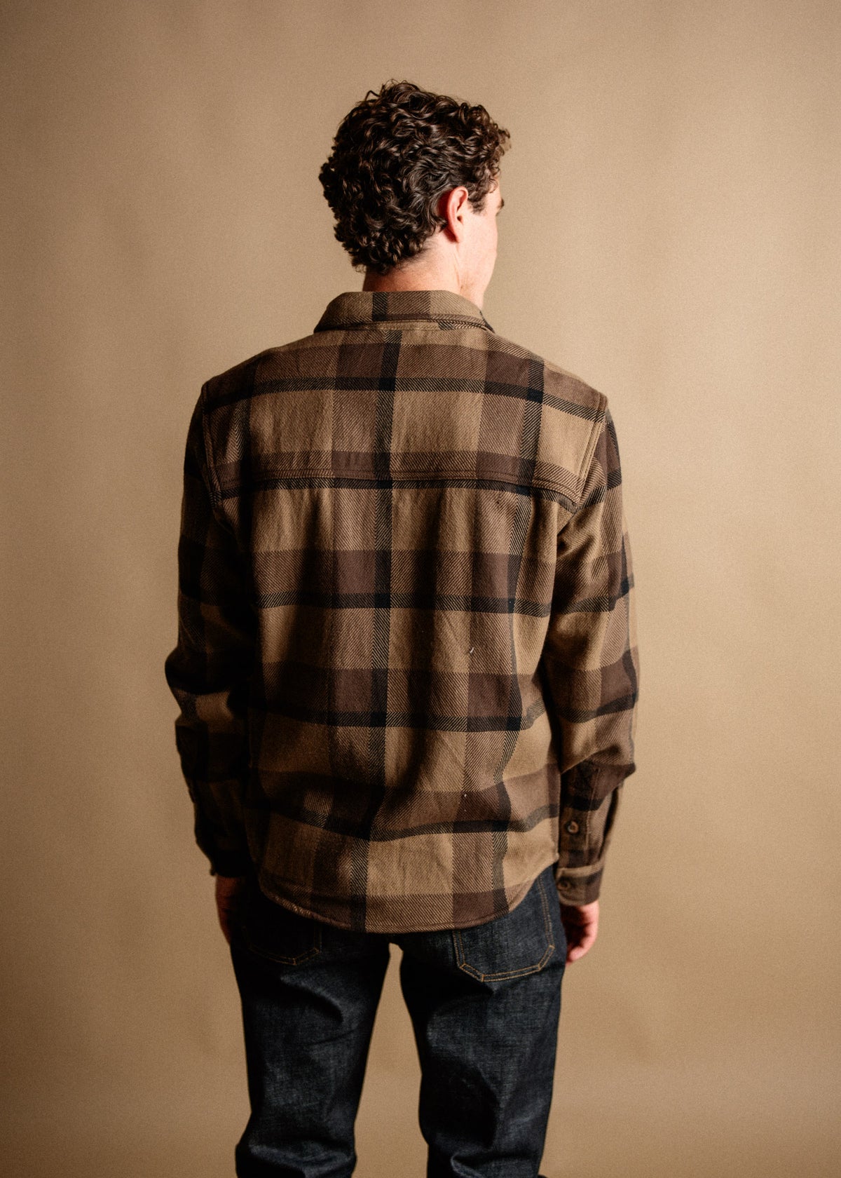 Man with curly hair in a brown plaid shirt and jeans, facing away against a beige background.