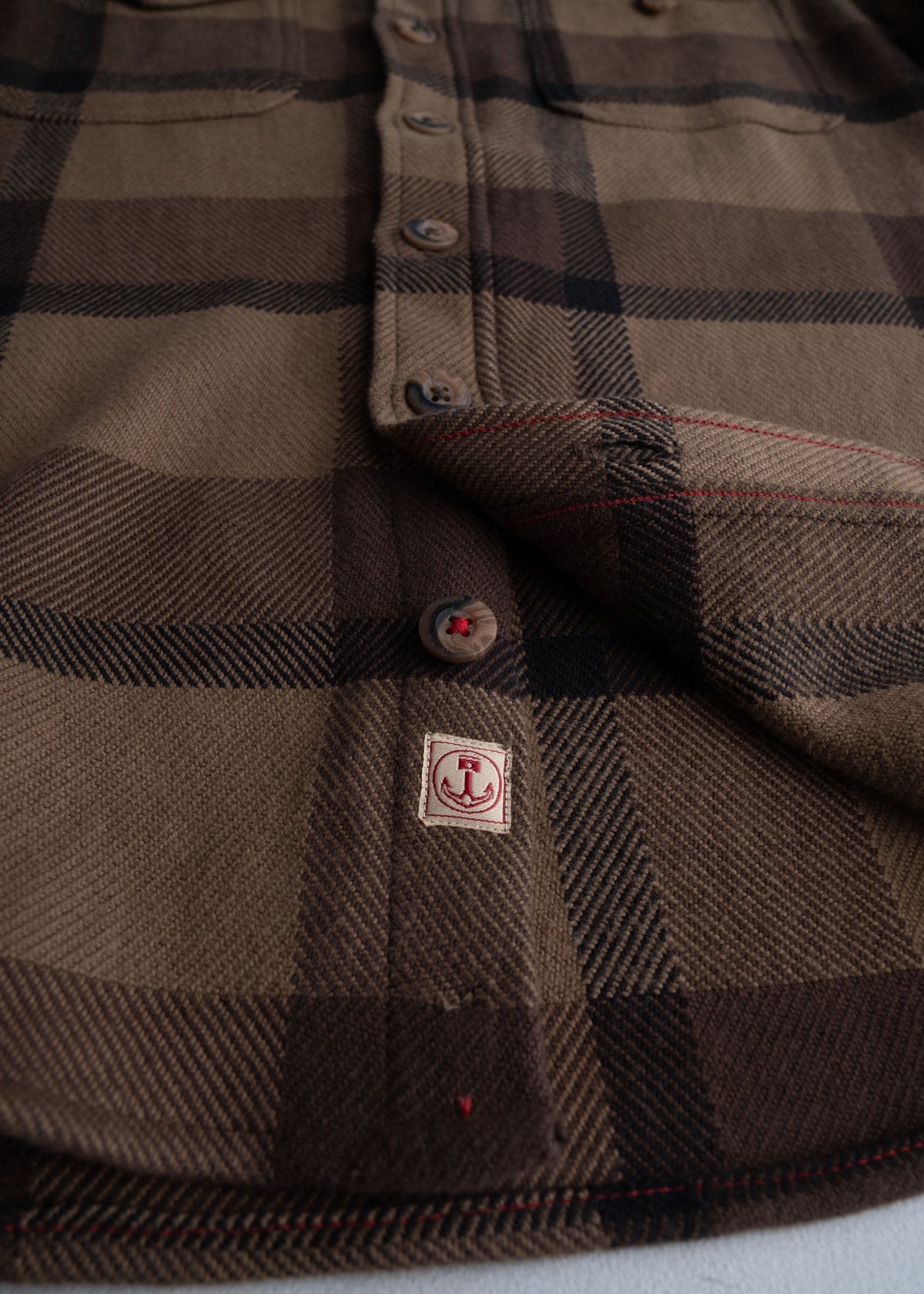 Close-up of a brown plaid shirt with buttons and an anchor patch, highlighting the fabric texture and design details.