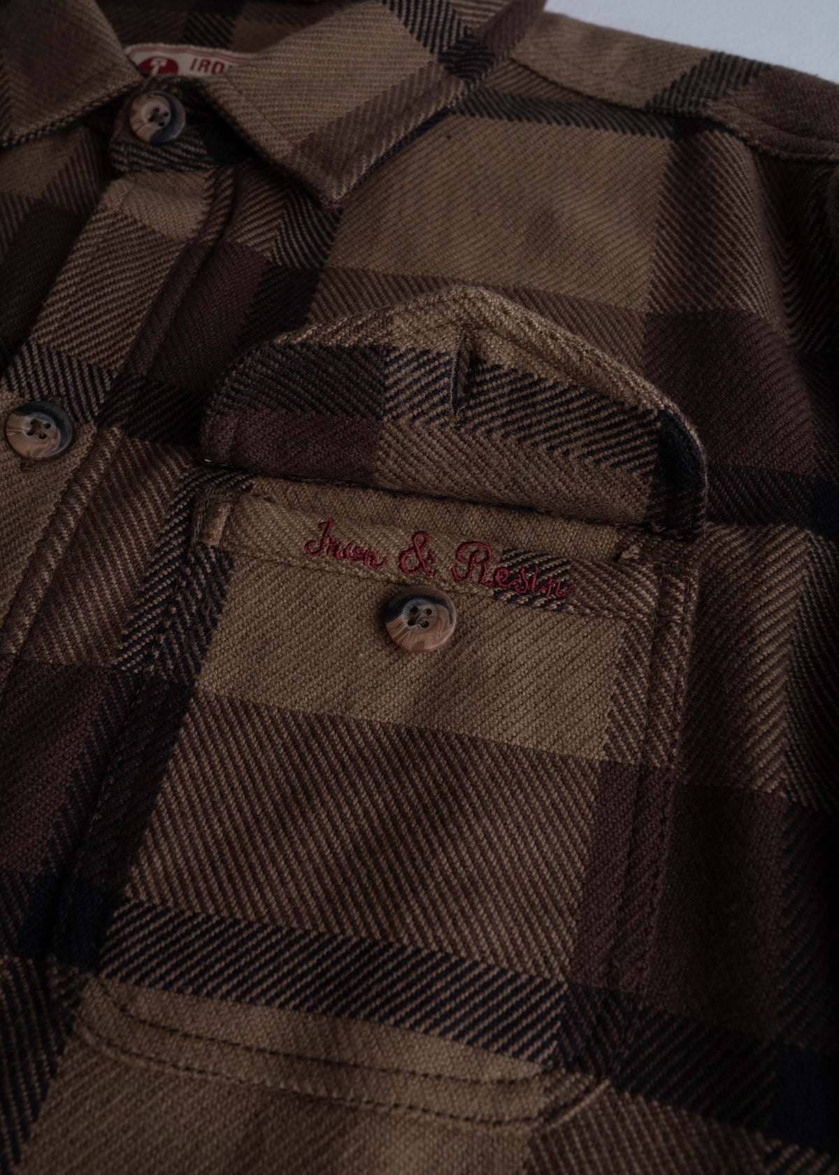 Close-up of a brown plaid shirt with "Iron & Resin" embroidered above the pocket, showcasing detailed stitching and buttons.
