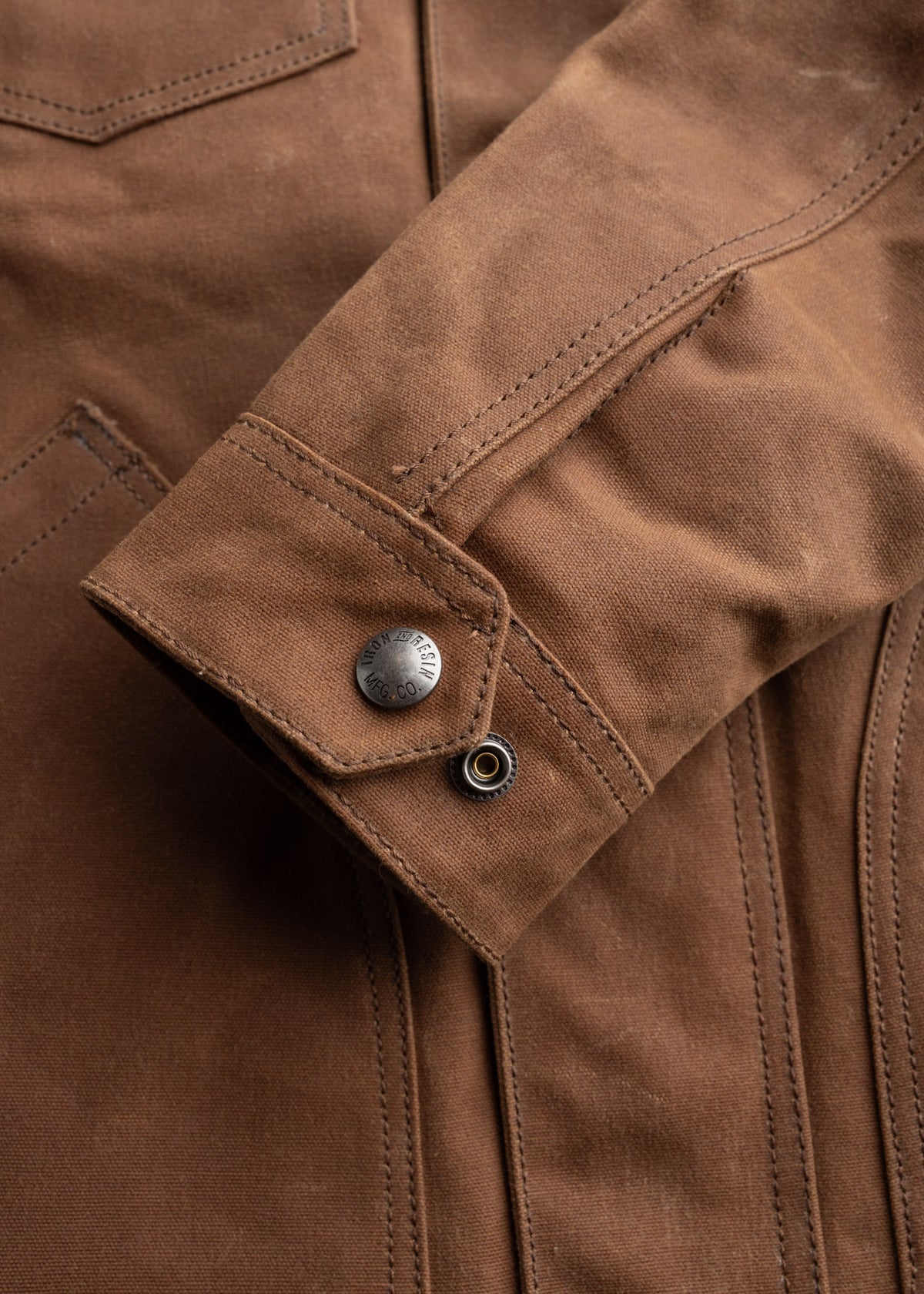 Close-up of a brown jacket sleeve with detailed stitching and a metal button, showcasing craftsmanship and texture.