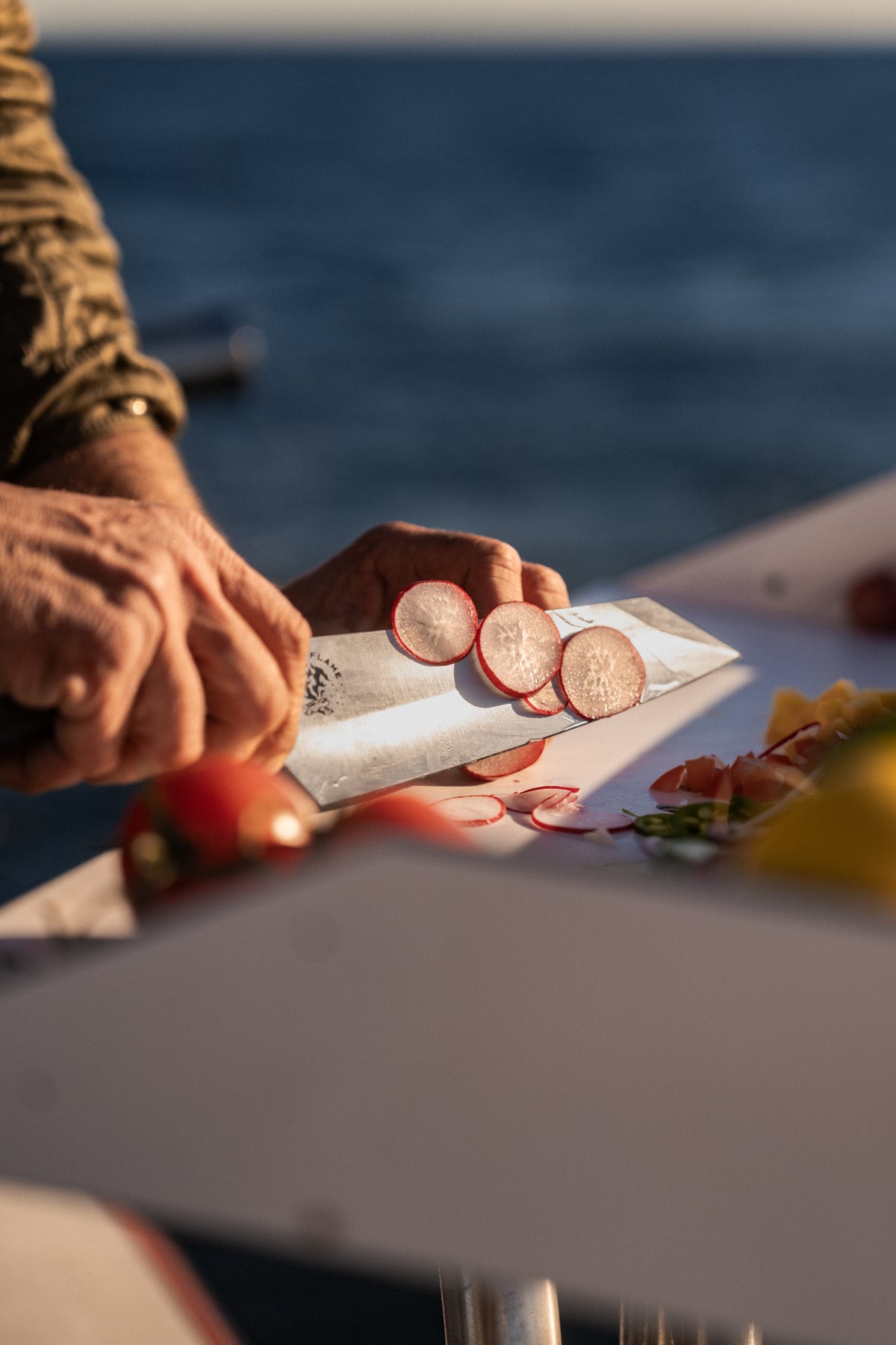 A person slices radishes with a knife on a boat, with fresh vegetables nearby and the ocean in the background.