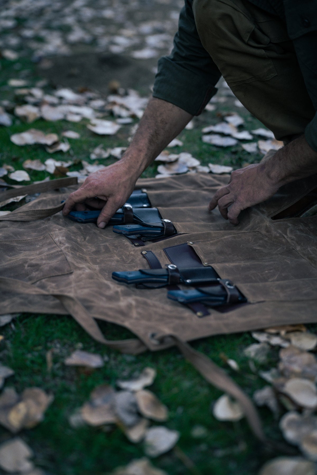 A person arranges knives on a brown canvas roll outdoors, surrounded by fallen leaves, highlighting a rustic, natural setting.