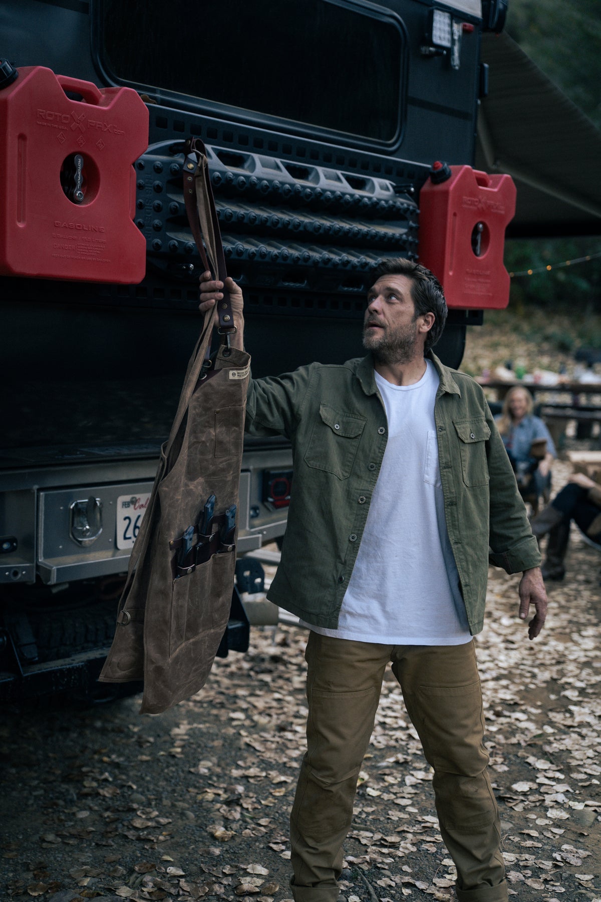 Man in green jacket holds a tool apron near a rugged vehicle with red fuel cans, surrounded by fallen leaves and outdoor gear.