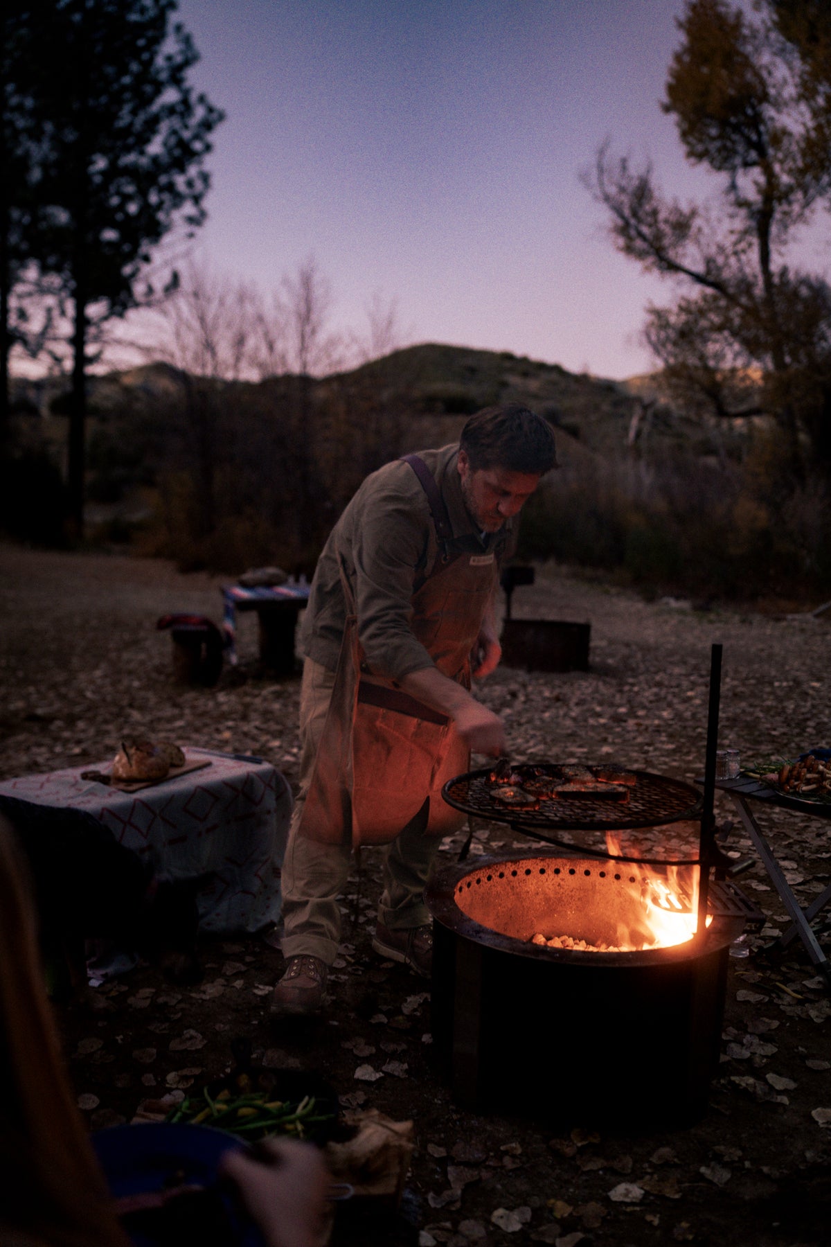 A person in an apron cooks over a campfire grill at dusk, surrounded by trees and hills, creating a cozy outdoor setting.
