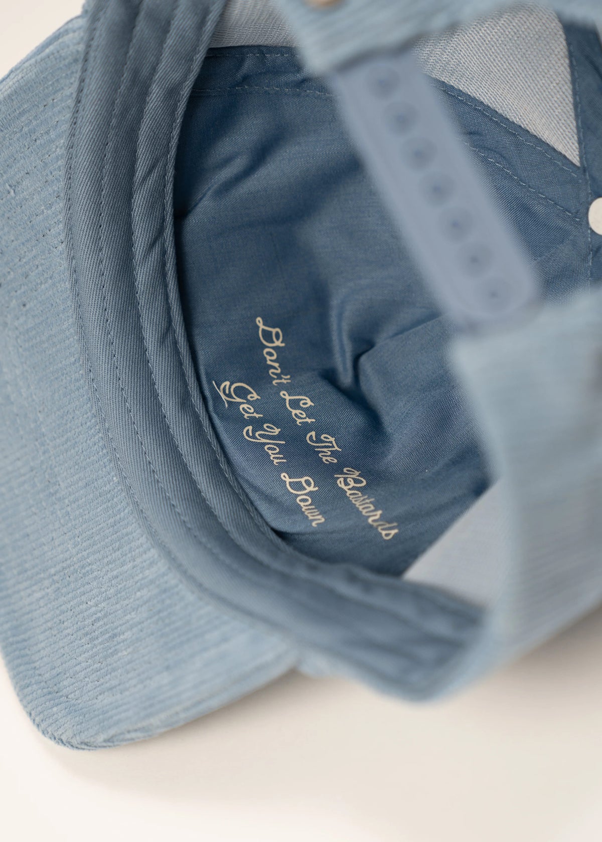 Blue cap interior with embroidered message: "Don't Let The Bastards Get You Down," offering a motivational reminder.