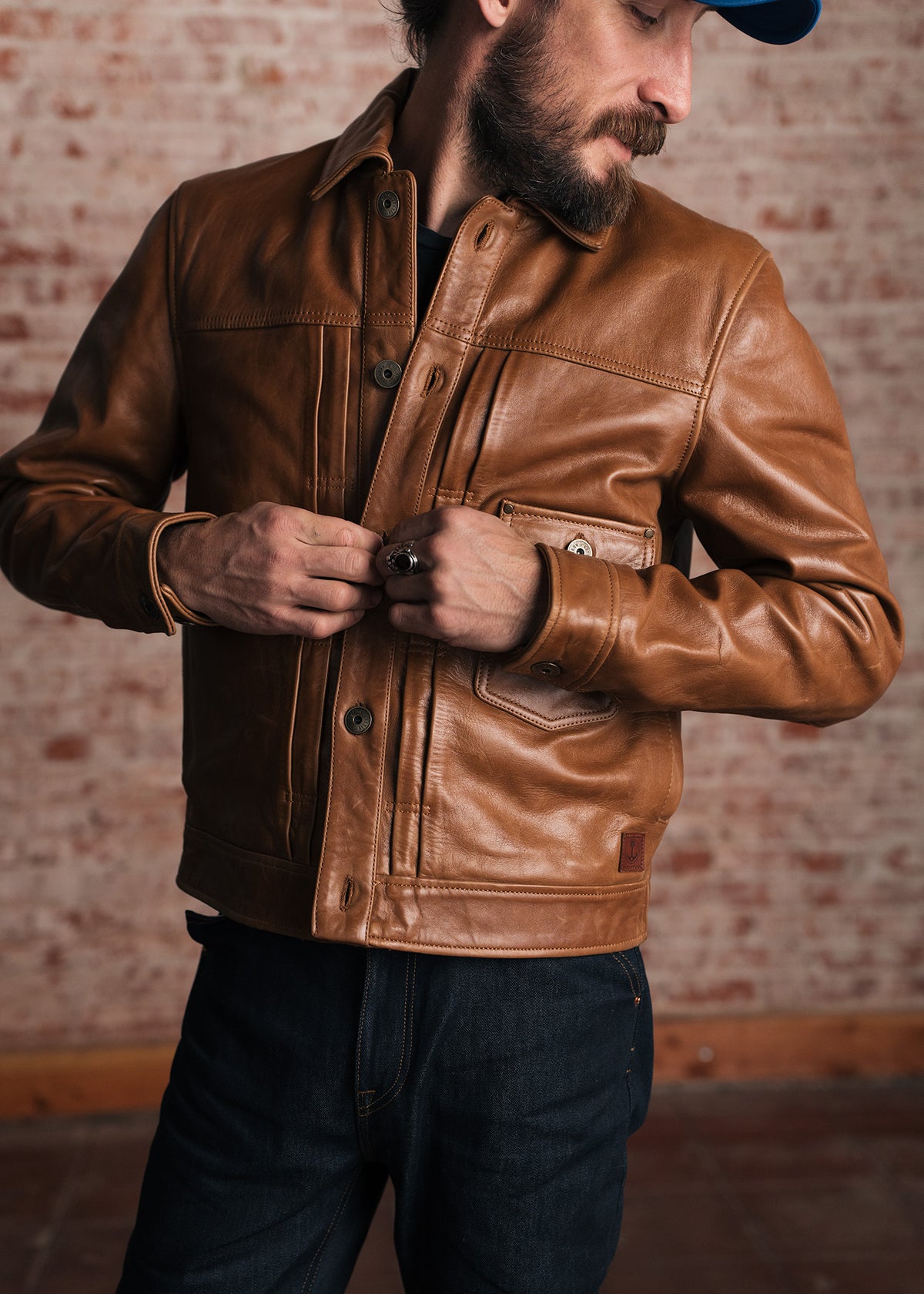 Man in a brown leather jacket adjusts buttons, wearing a blue cap, standing against a brick wall background.
