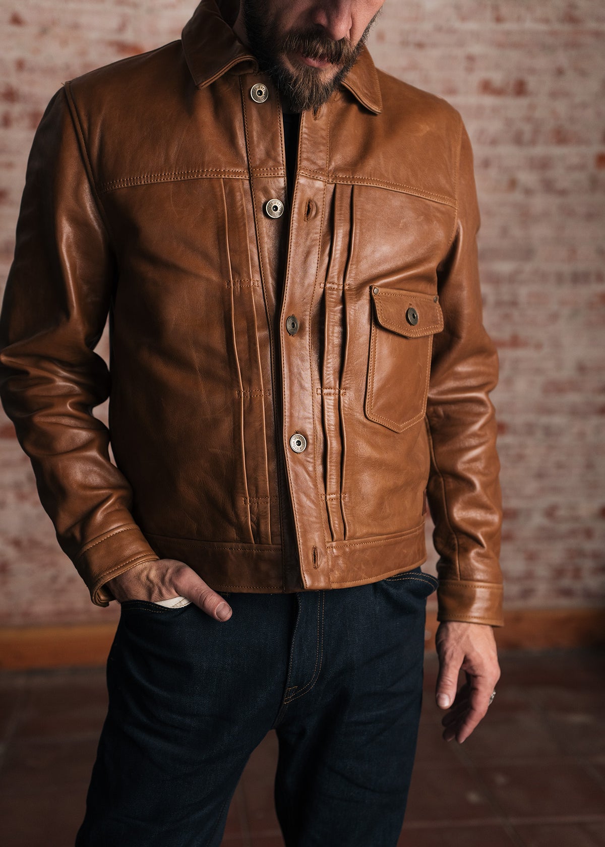 Man in brown leather jacket and jeans stands against a brick wall, hands in pockets, showcasing casual style and texture.