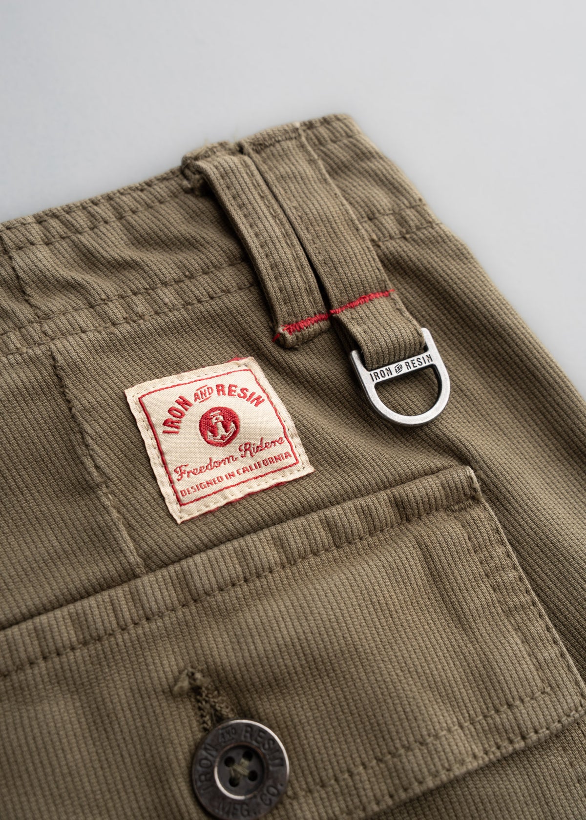 Close-up of khaki pants with Iron and Resin label, button, and D-ring detail, highlighting craftsmanship and rugged style.