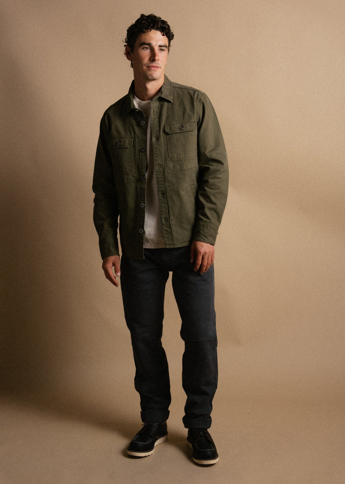 Man in green jacket and dark jeans stands against a beige background, looking to the side with a relaxed expression.
