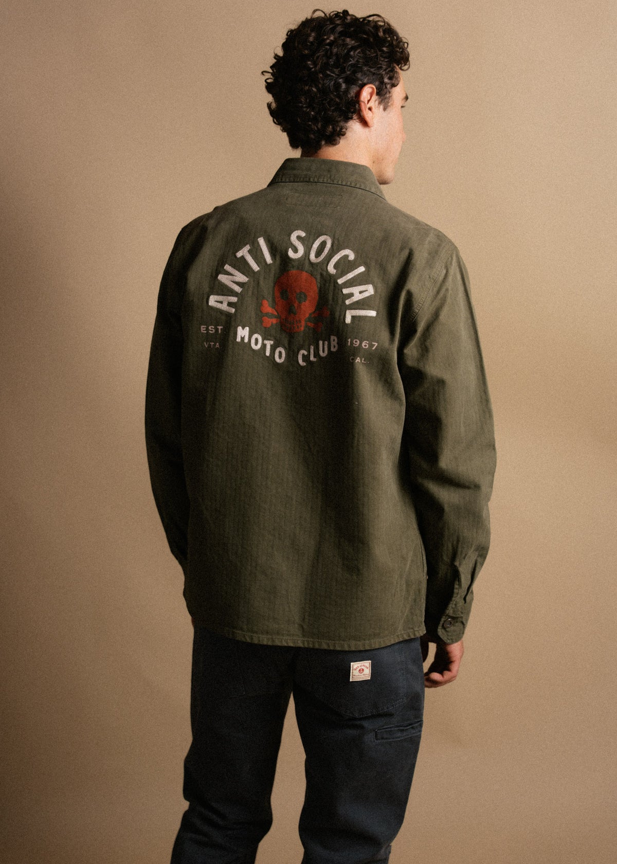 Man in olive jacket with "Anti Social Moto Club" and skull design on back, stands against a beige background, facing away.