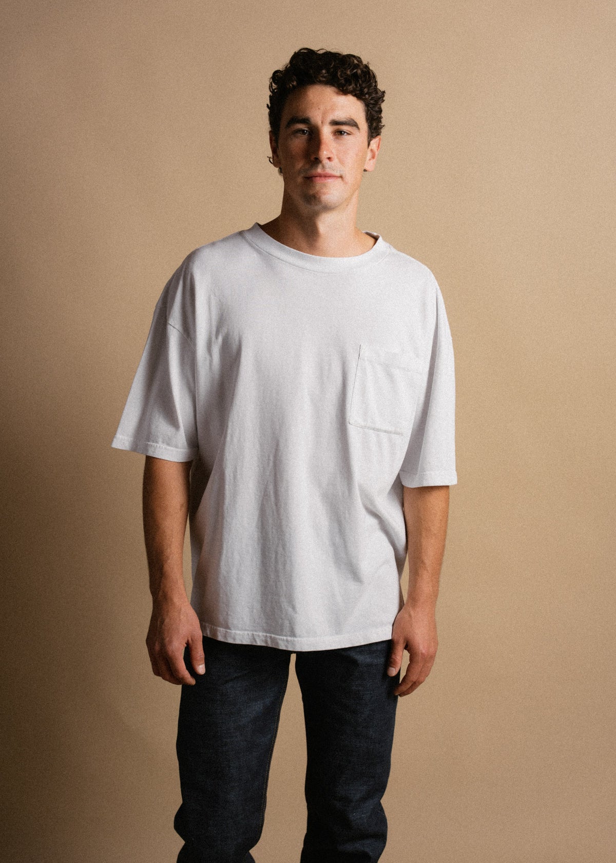 Man in a white t-shirt and jeans stands against a beige background, looking confidently at the camera with a neutral expression.