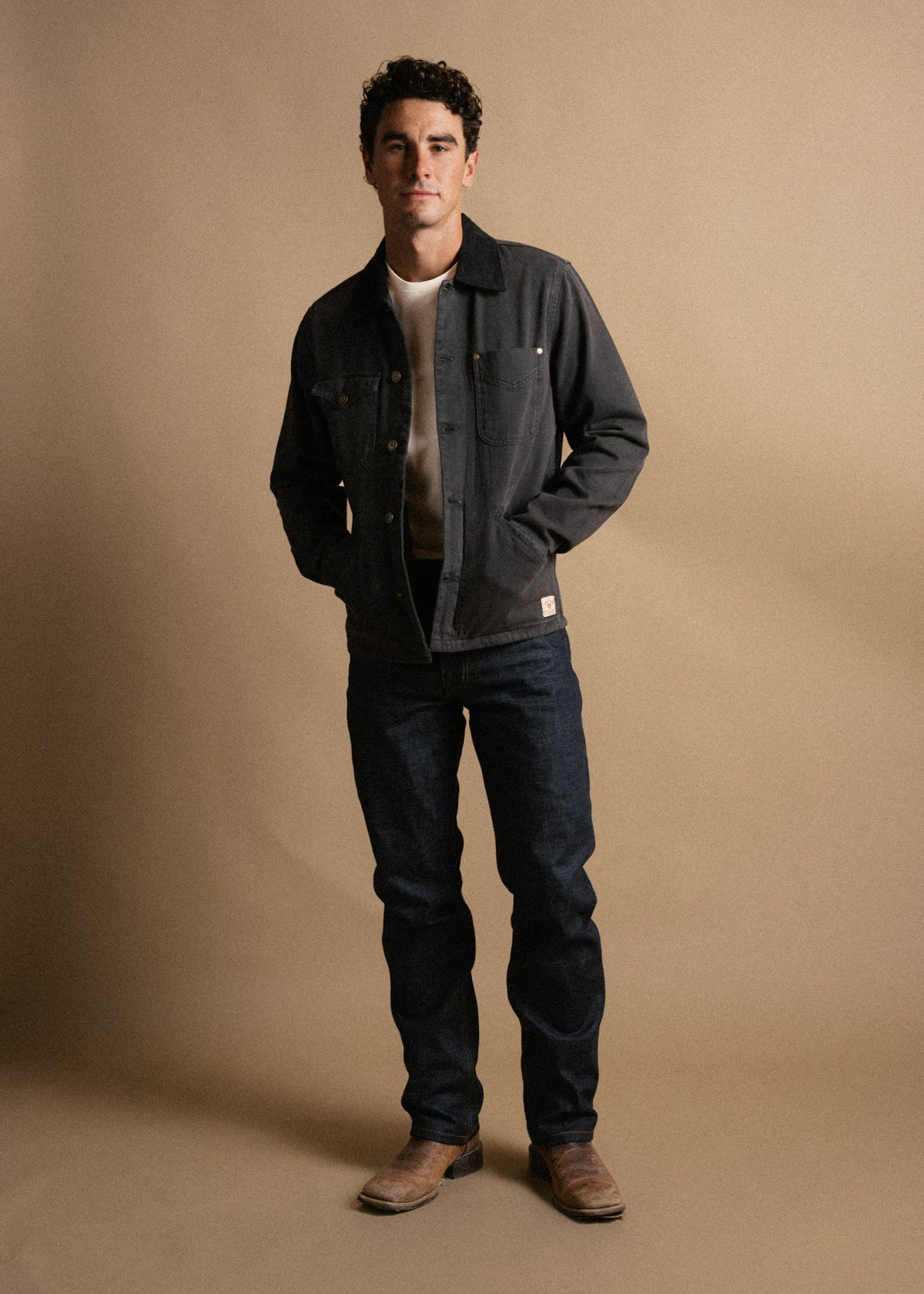 Man in denim jacket and jeans stands confidently against a beige background, hands in pockets, wearing brown boots.