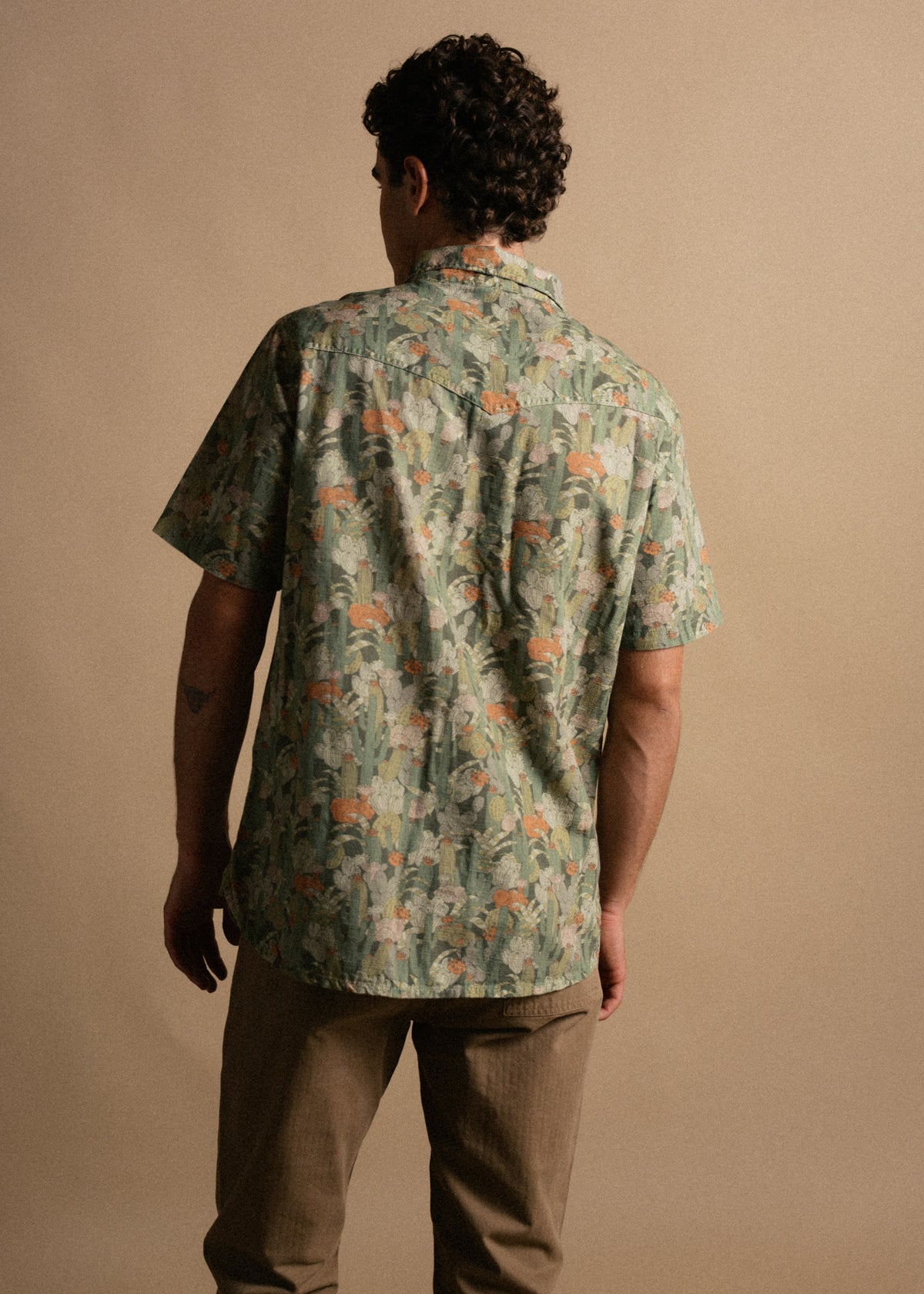 Man in a floral short-sleeve shirt and khaki pants stands with his back to the camera against a plain beige background.