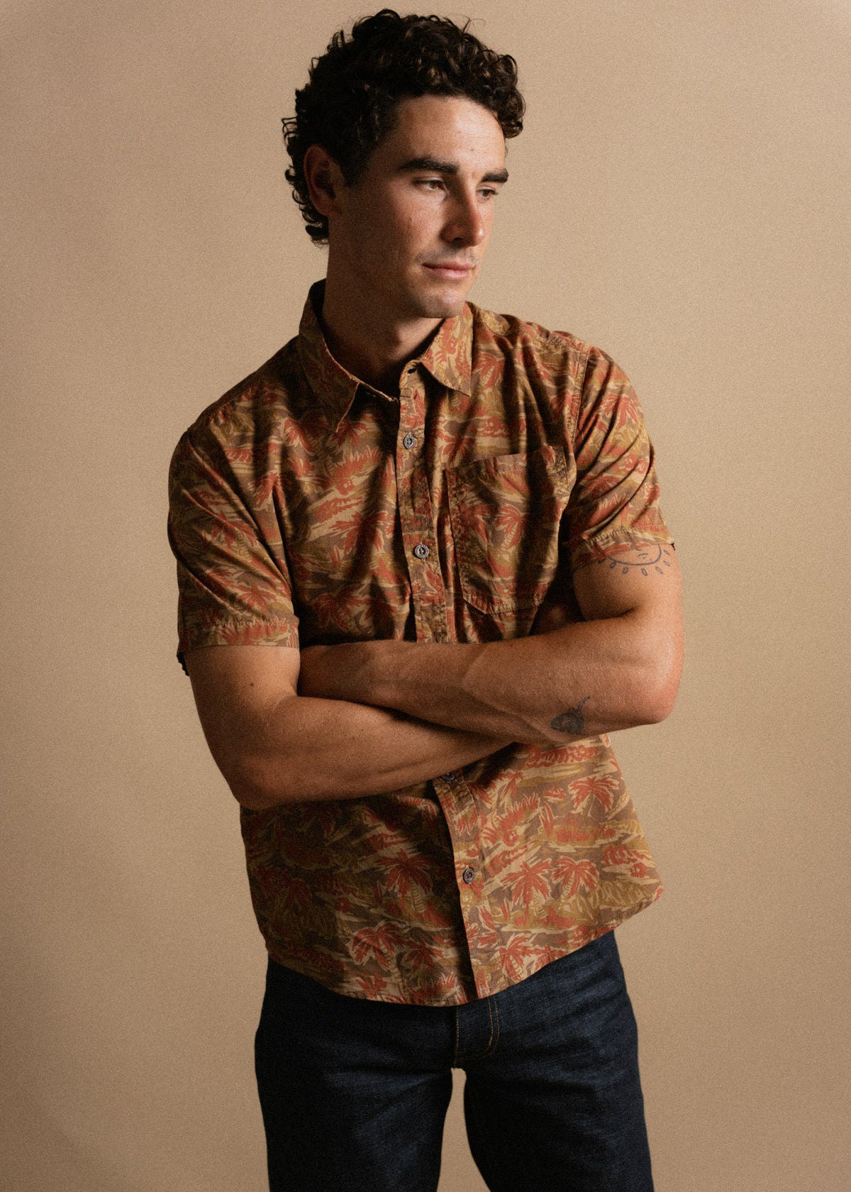 Man in a patterned shirt and jeans stands with arms crossed against a beige background, looking to the side thoughtfully.
