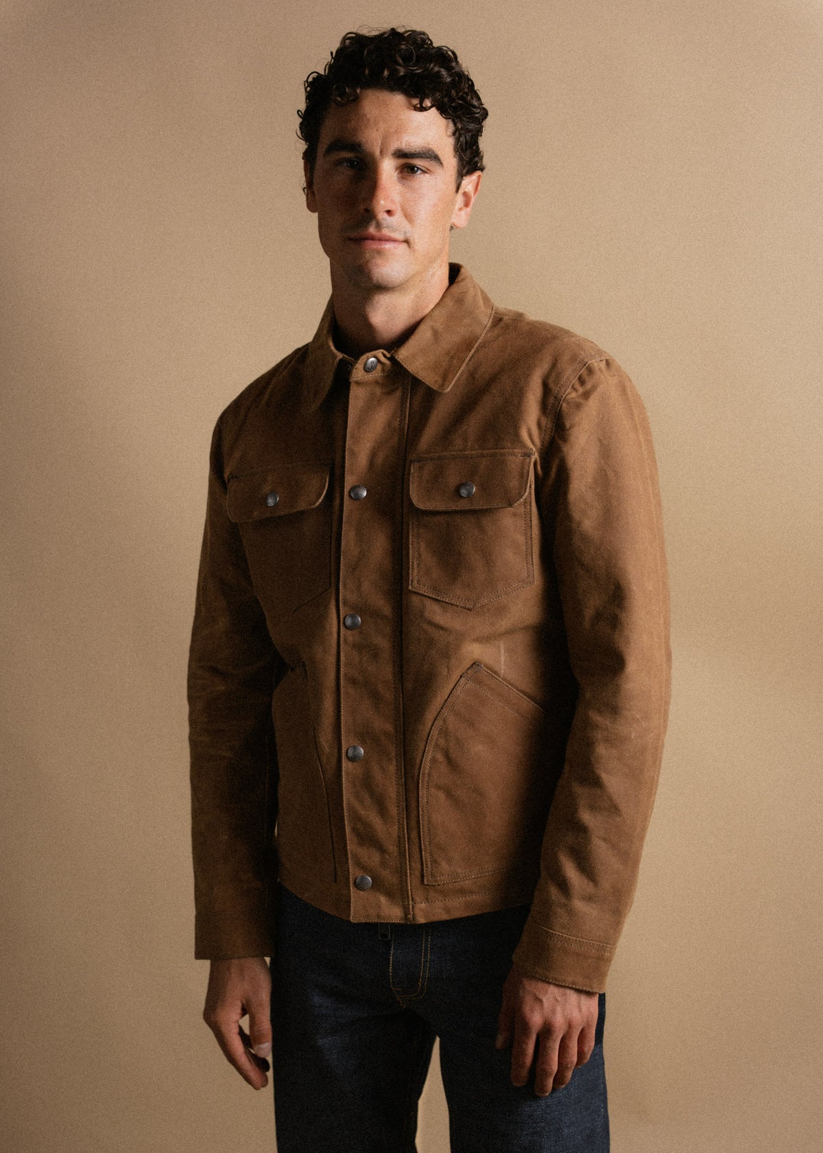 Man in a brown jacket with buttoned pockets stands against a beige background, looking confidently at the camera.