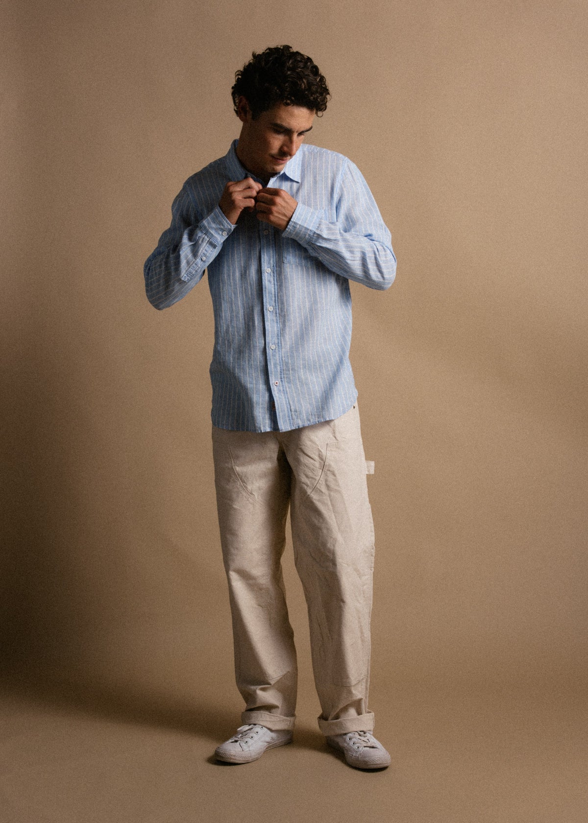 Man in blue striped shirt and beige pants adjusts collar, standing against a plain brown background, wearing white sneakers.