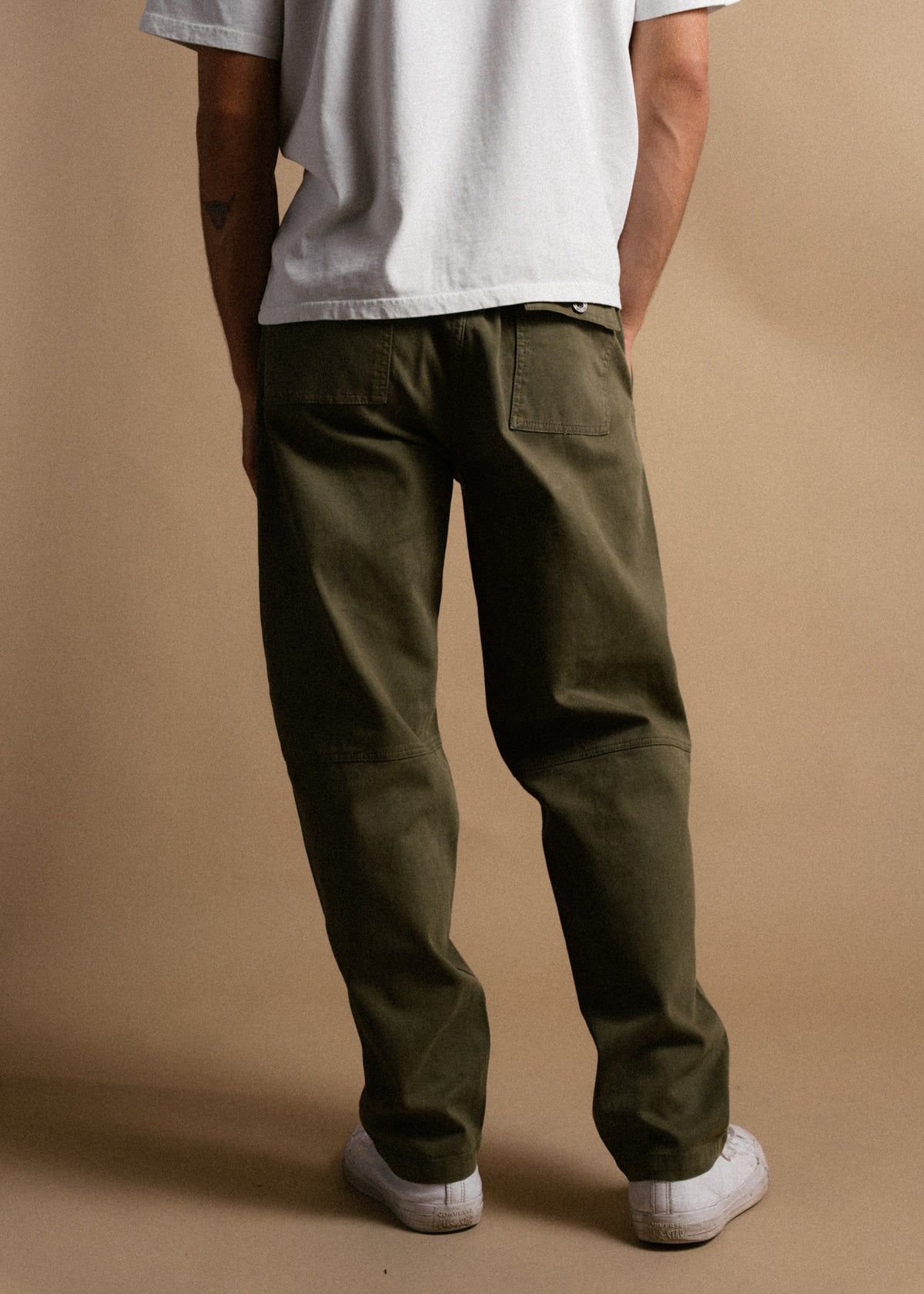 Man in olive pants and white sneakers stands against a beige background, wearing a white shirt, facing away.