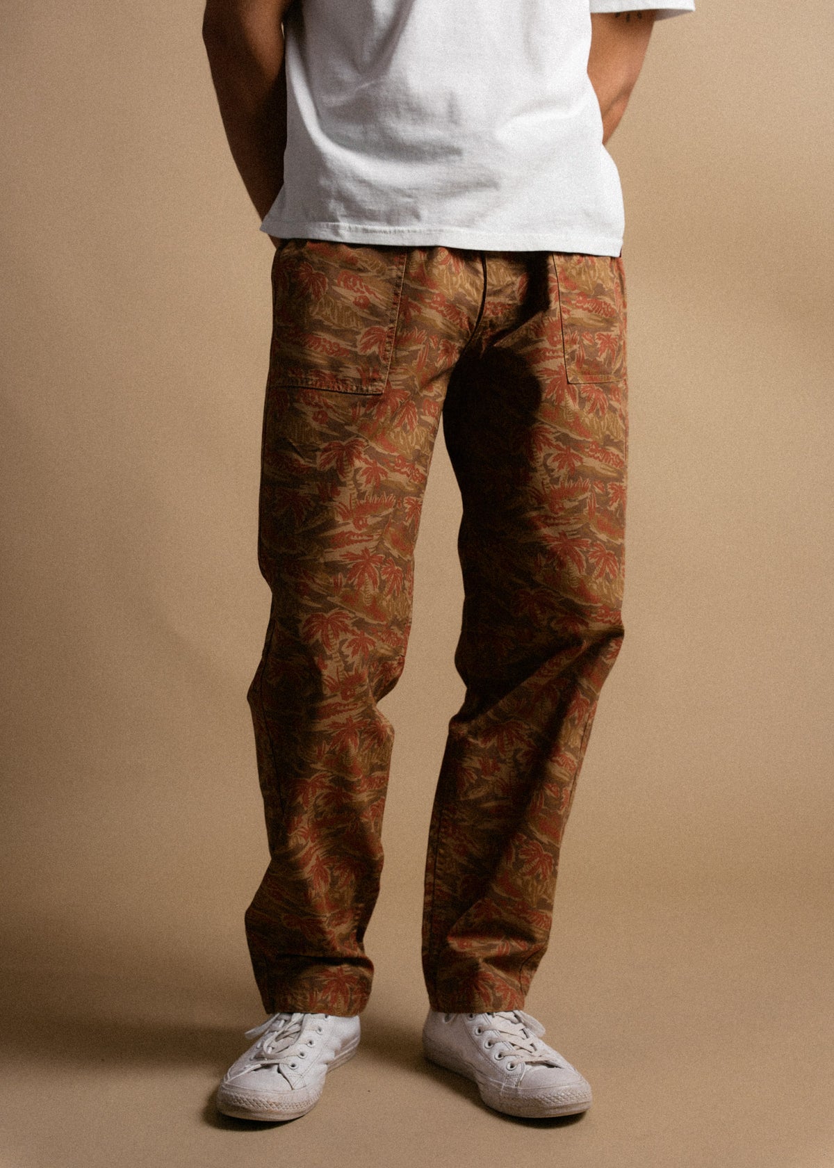 A person in a white shirt and patterned brown pants stands against a beige background, wearing white sneakers.