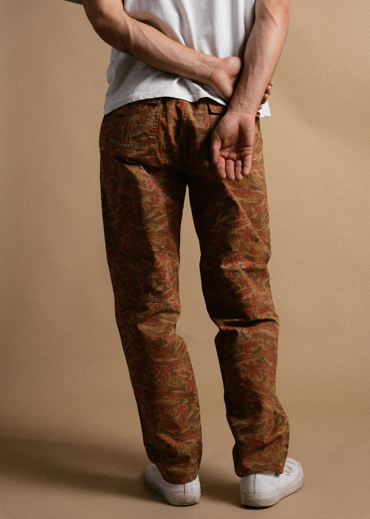 Man in white shirt and patterned pants stands with hands behind back, facing away on beige background. White sneakers visible.