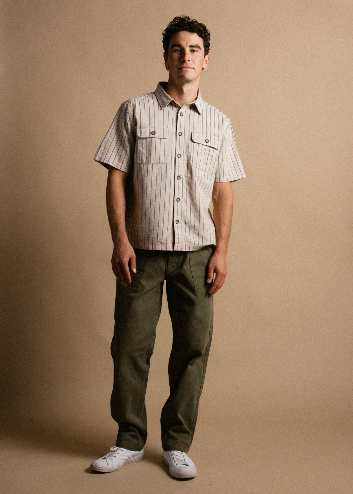 Man in striped shirt, olive pants, and white sneakers stands against a beige backdrop, exuding casual style and confidence.