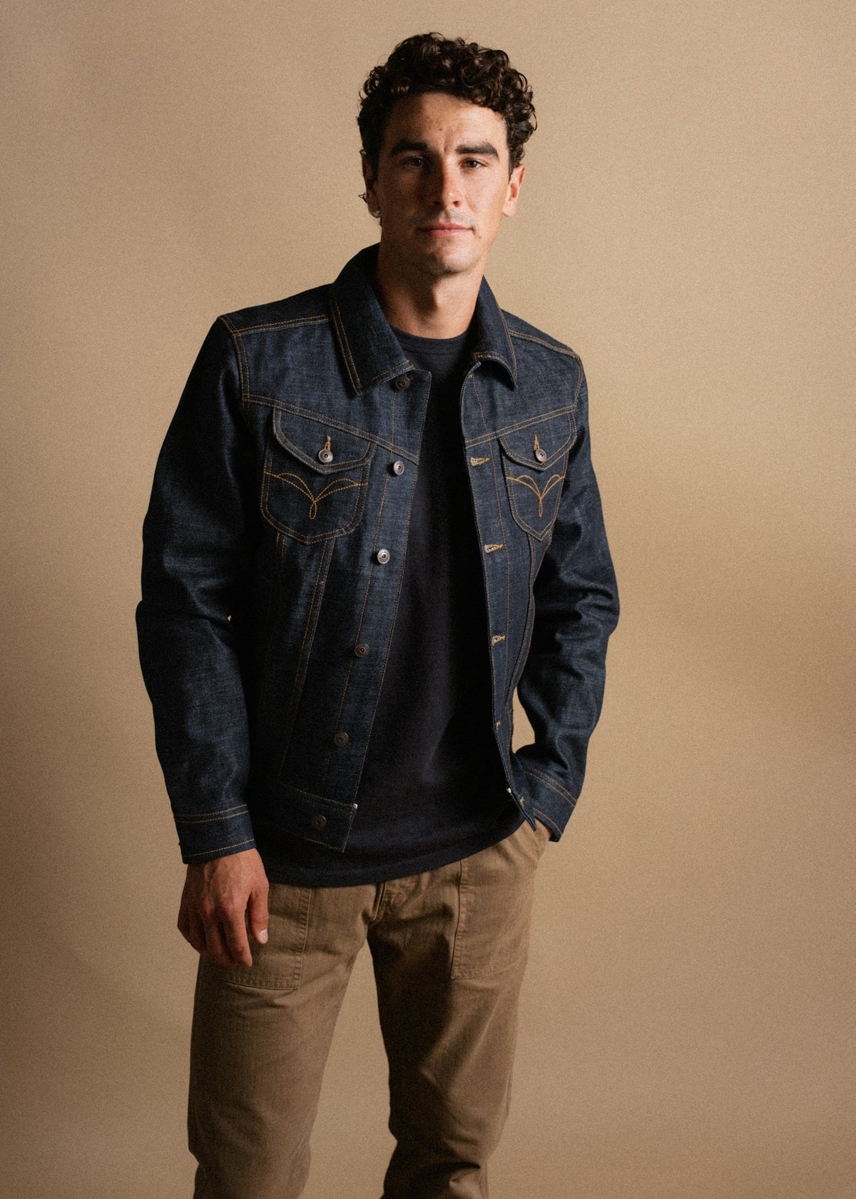 Man in a denim jacket and khaki pants stands against a beige background, exuding a casual and confident style.