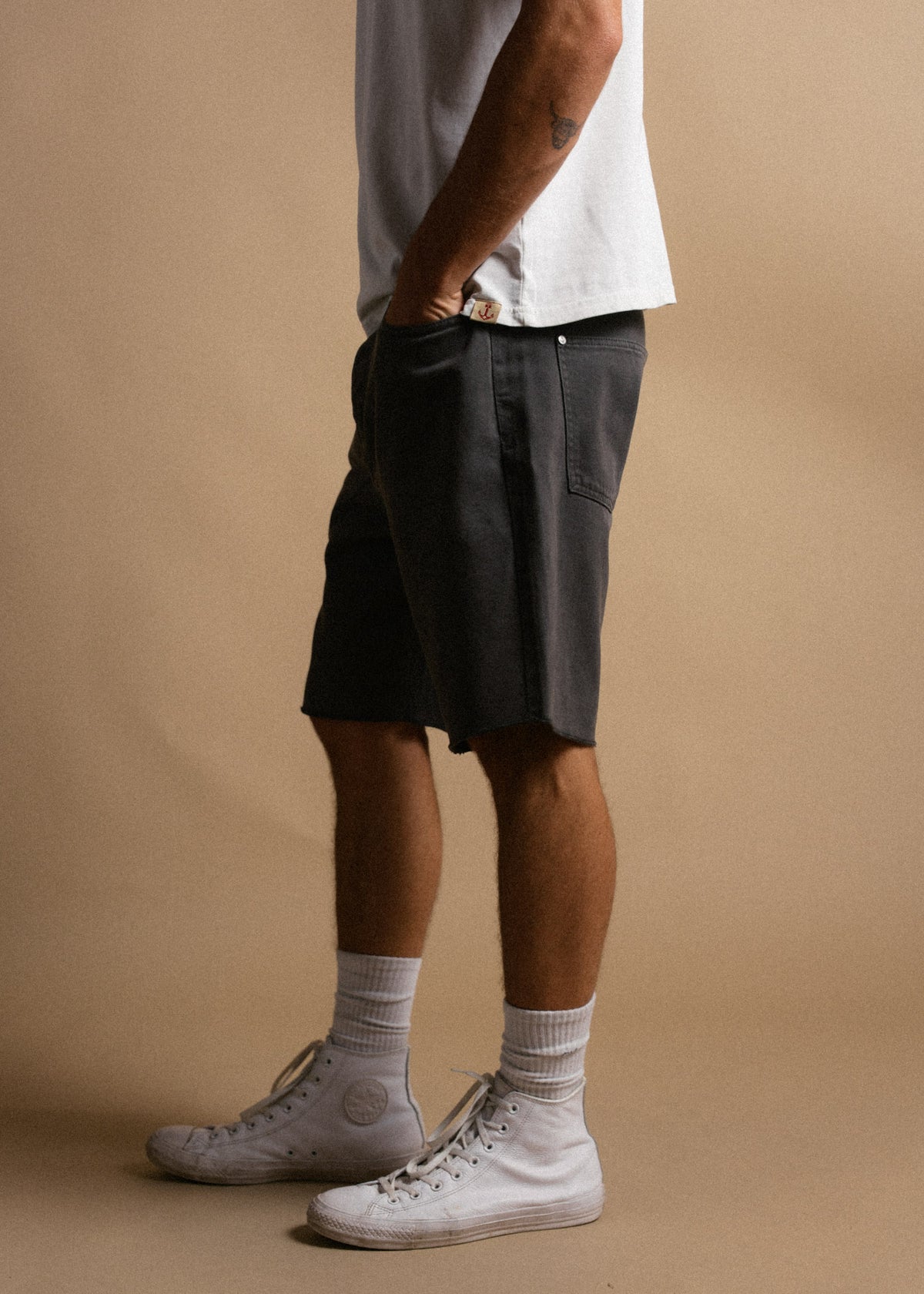 Man in white shirt and black shorts stands sideways, hands in pockets, wearing white sneakers and socks on a beige background.