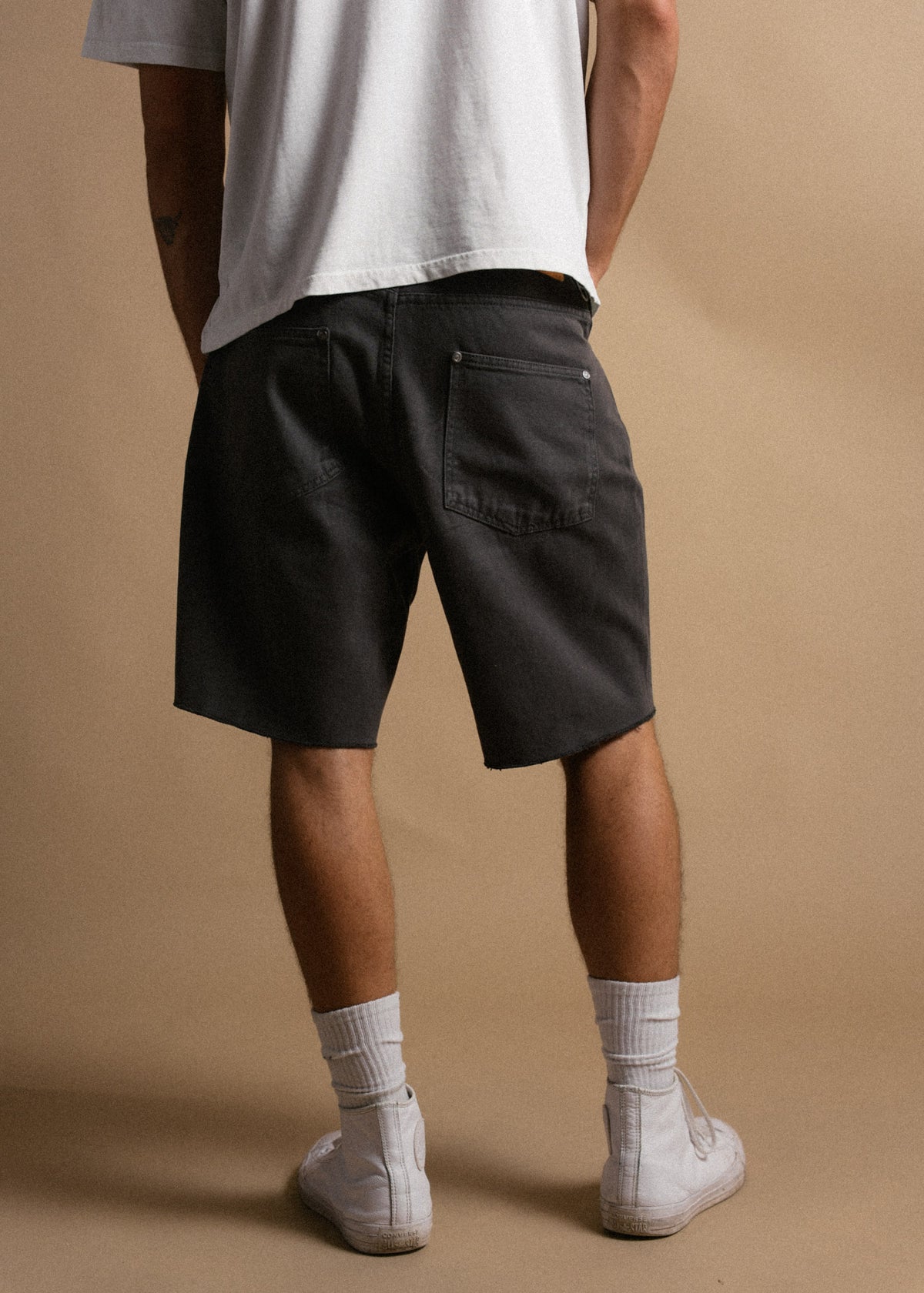 Man in white shirt, black shorts, and white sneakers stands against a beige background, hands in pockets, facing away.