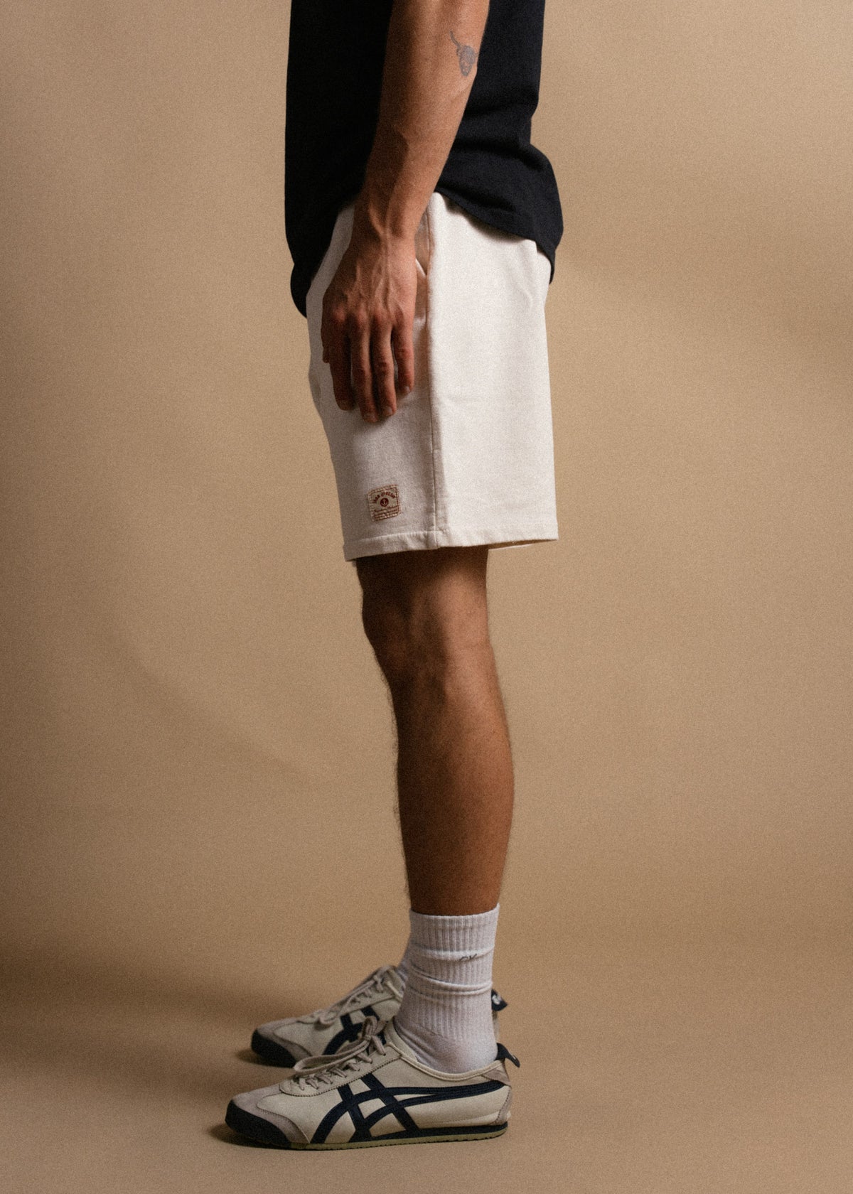 Man in white shorts and sneakers stands against a beige background, showcasing casual style with a black shirt and socks.