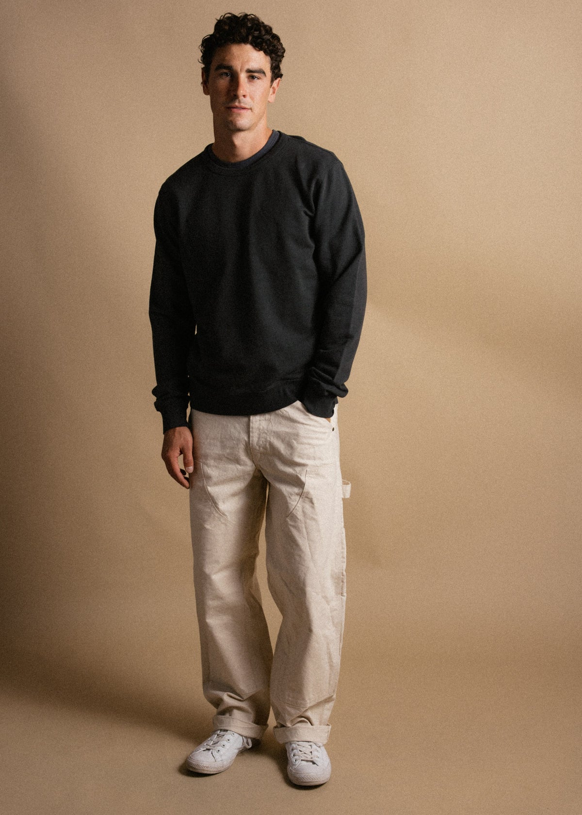 Man in a black sweater and beige pants stands against a neutral backdrop, exuding casual style with a relaxed posture.