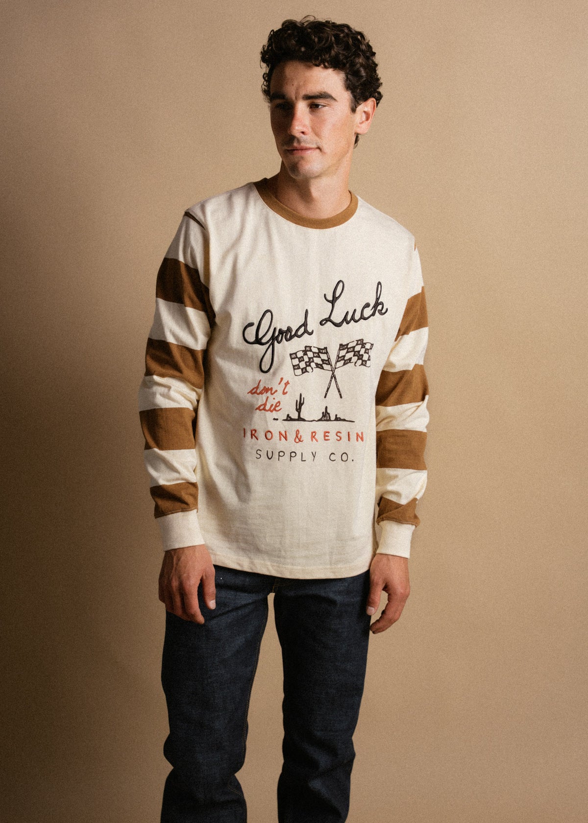 Man in a striped long-sleeve shirt with "Good Luck" text, standing against a beige background, wearing dark jeans.