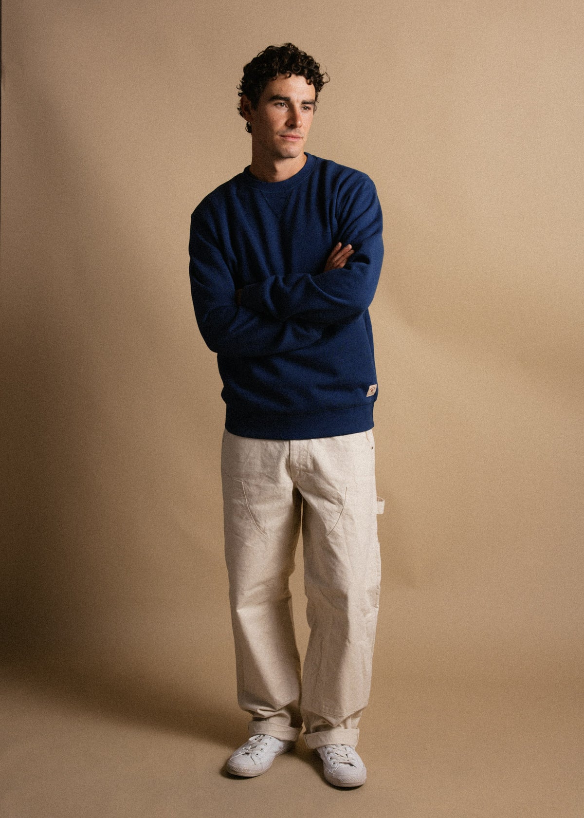 Man in blue sweater and beige pants stands against a tan background, arms crossed, looking to the side with a thoughtful expression.