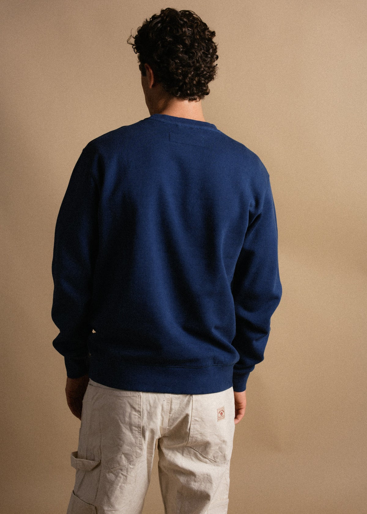Man in a blue sweatshirt and beige cargo pants stands with his back to the camera against a neutral background.