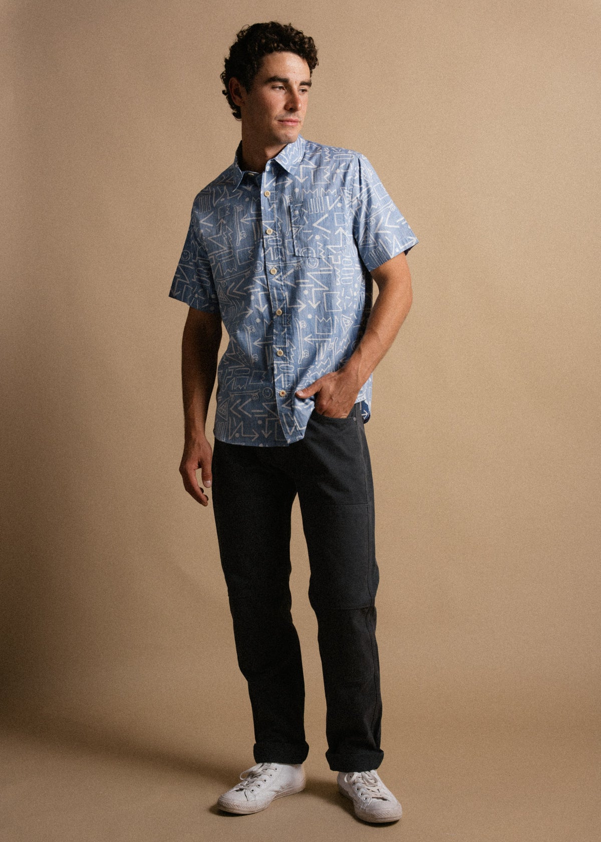 Man in a blue patterned shirt and black pants stands against a beige background, looking to the side with a relaxed posture.